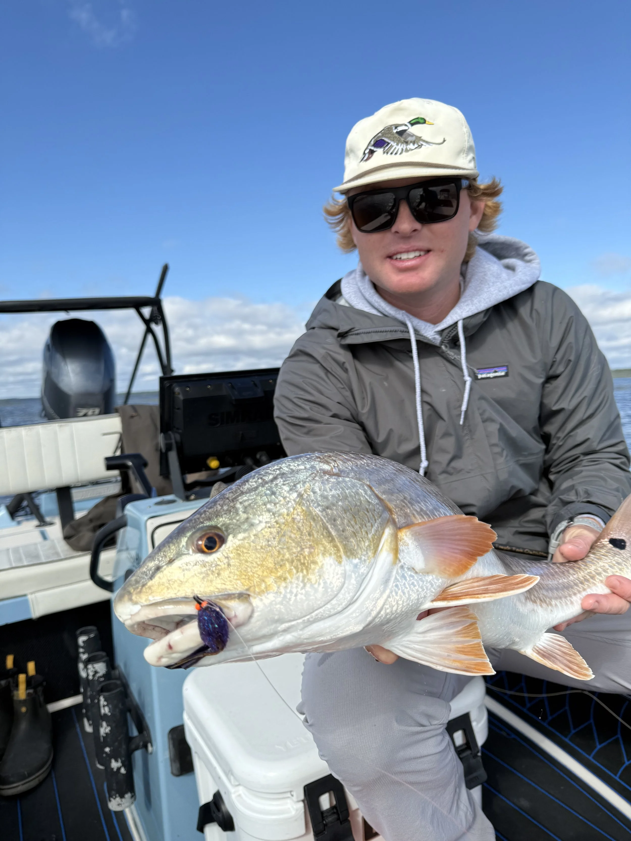 A young man on a boat holding a large fish with a purple lure in its mouth, under a partly cloudy sky.