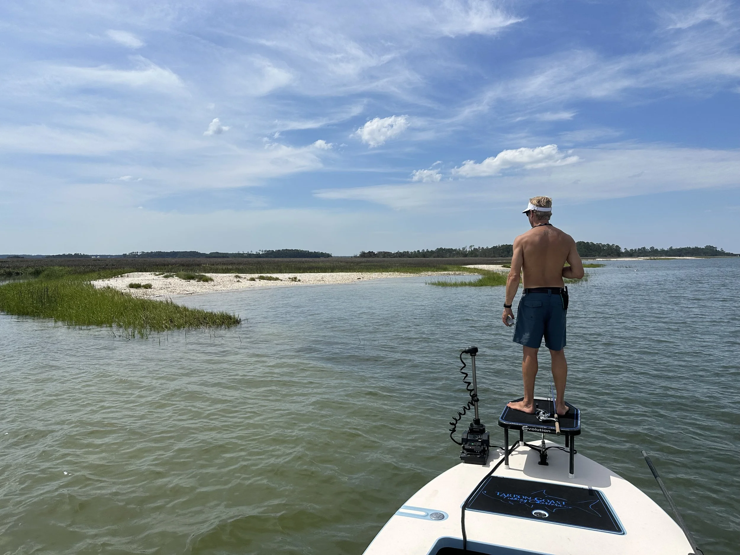 A man standing on a paddleboard, shirtless and wearing a cap, enjoying a scenic view of a waterway with grass and sandy banks under a blue sky with clouds.