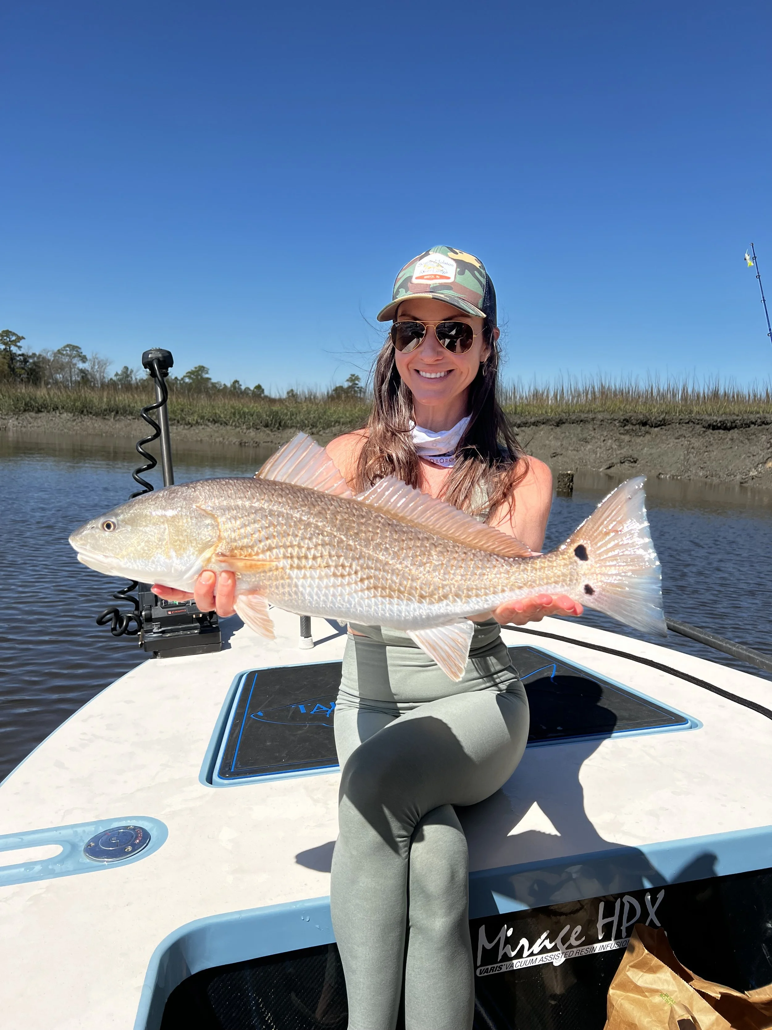Woman sitting on a boat holding a large fish, wearing sunglasses, a cap, and athletic clothing, with water and marshland in the background under clear blue skies.