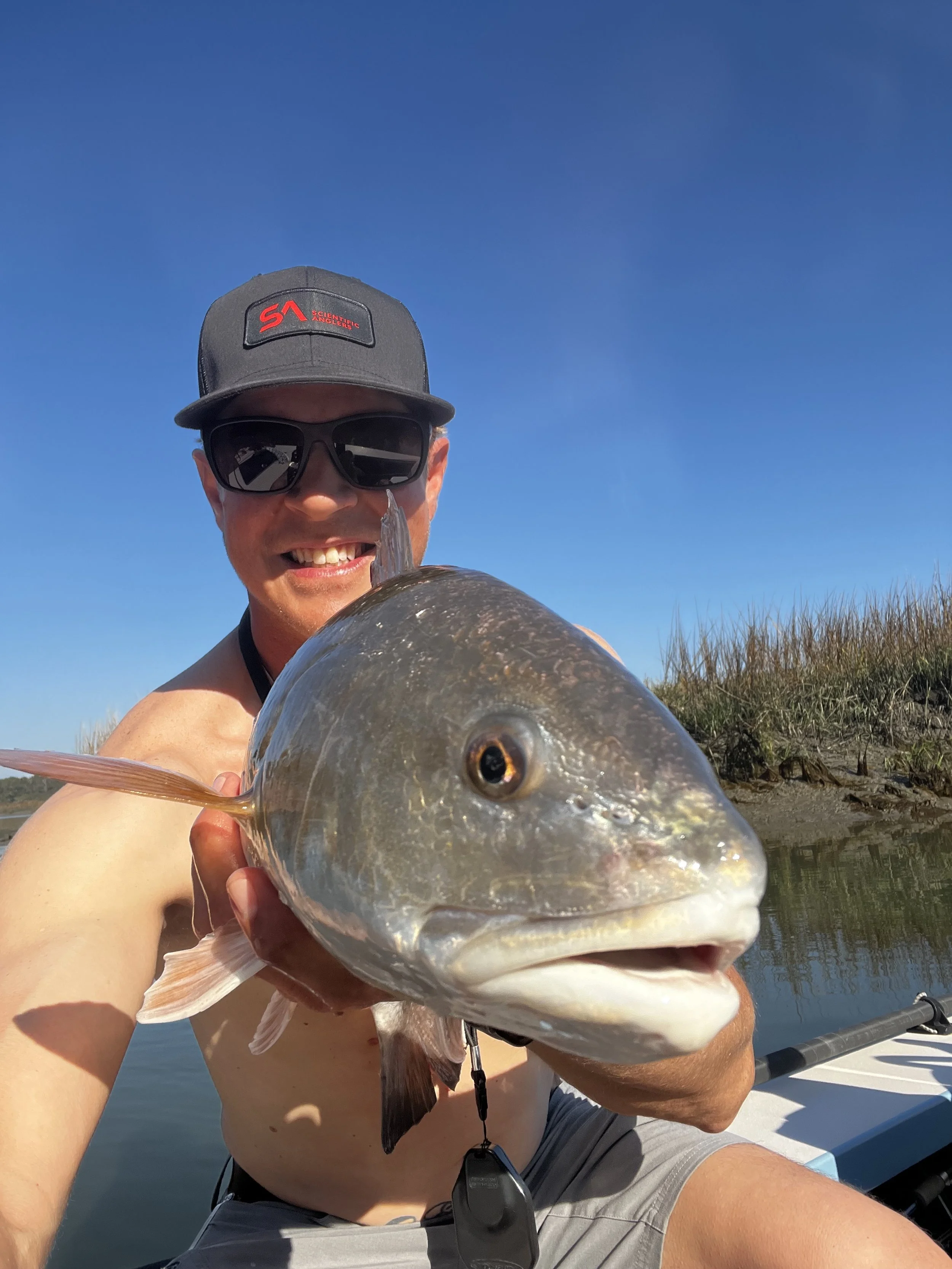 Man holding a large fish on a boat with blue sky and grassy shoreline in the background.
