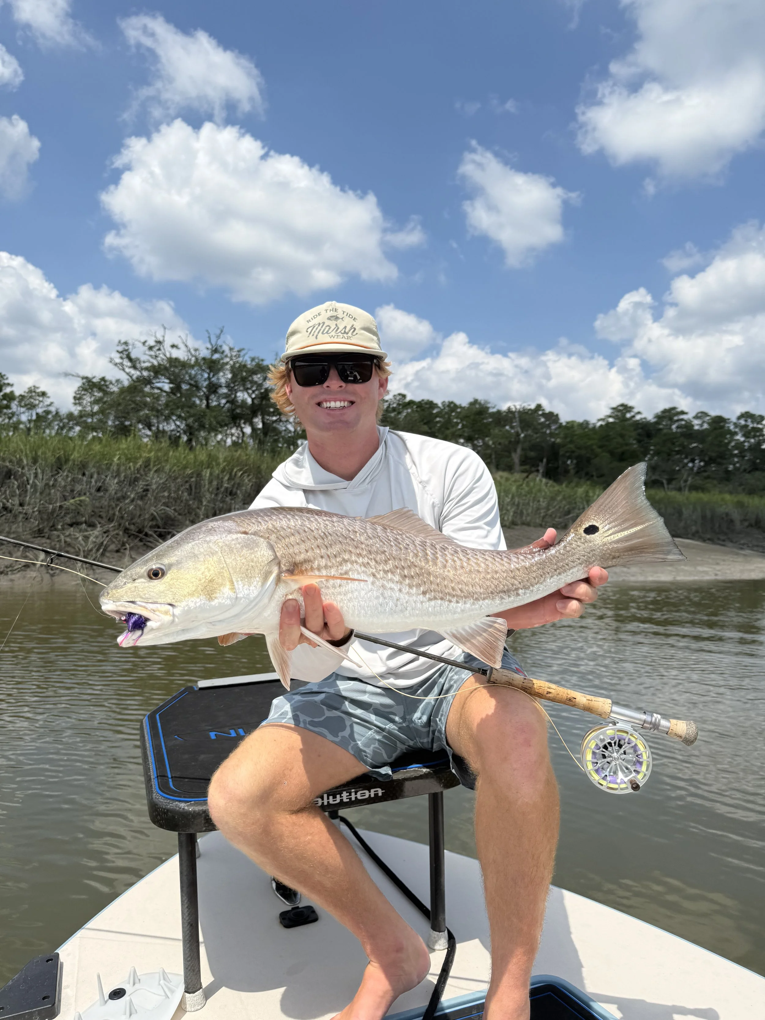 Young man sitting on a boat holding a large fish with a fishing rod in a river or lake with trees and a partly cloudy sky in the background.