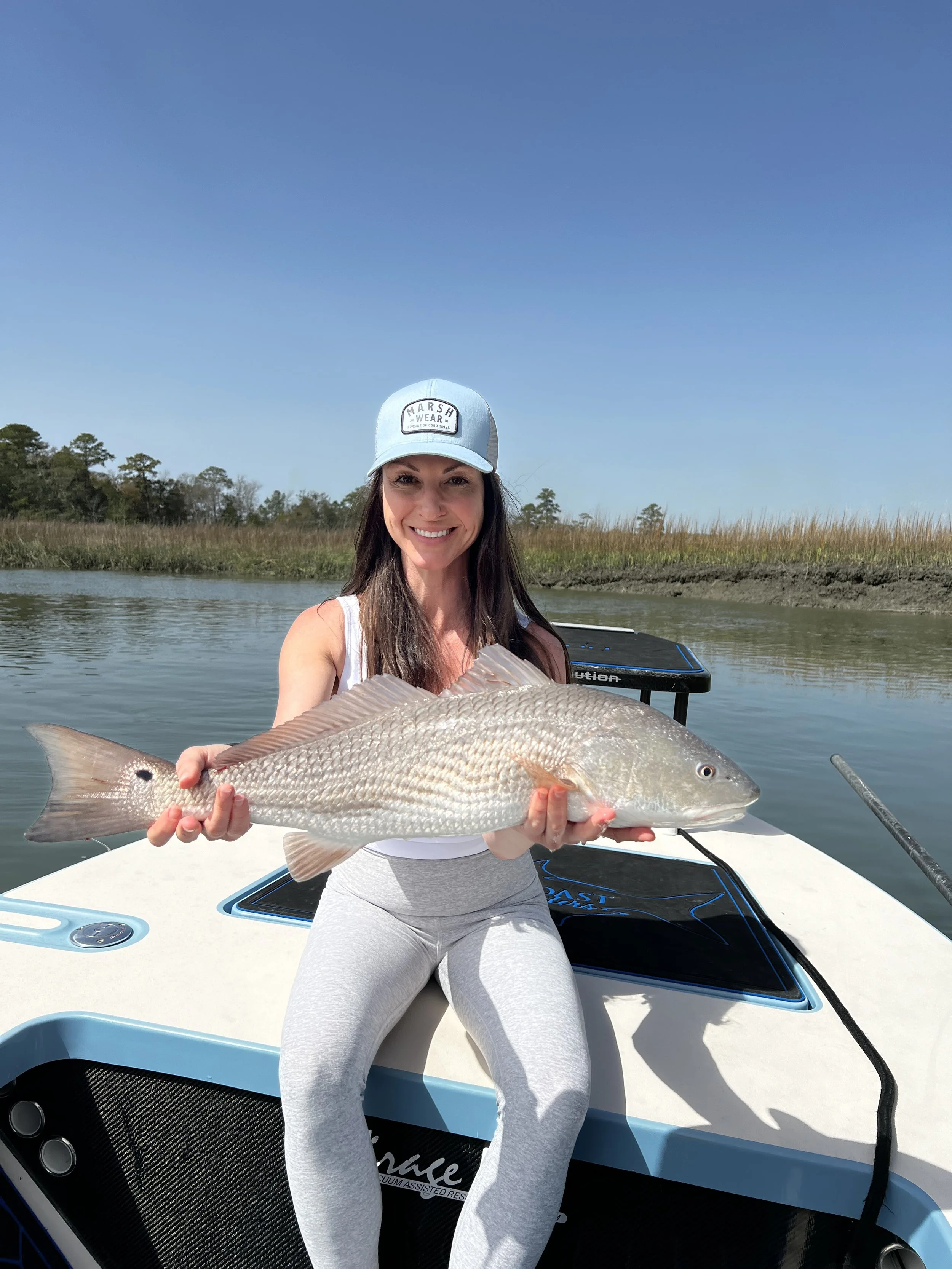 Woman sitting on boat holding a large fish, smiling, wearing a light blue baseball cap and gray leggings, with water and marsh grass in the background.
