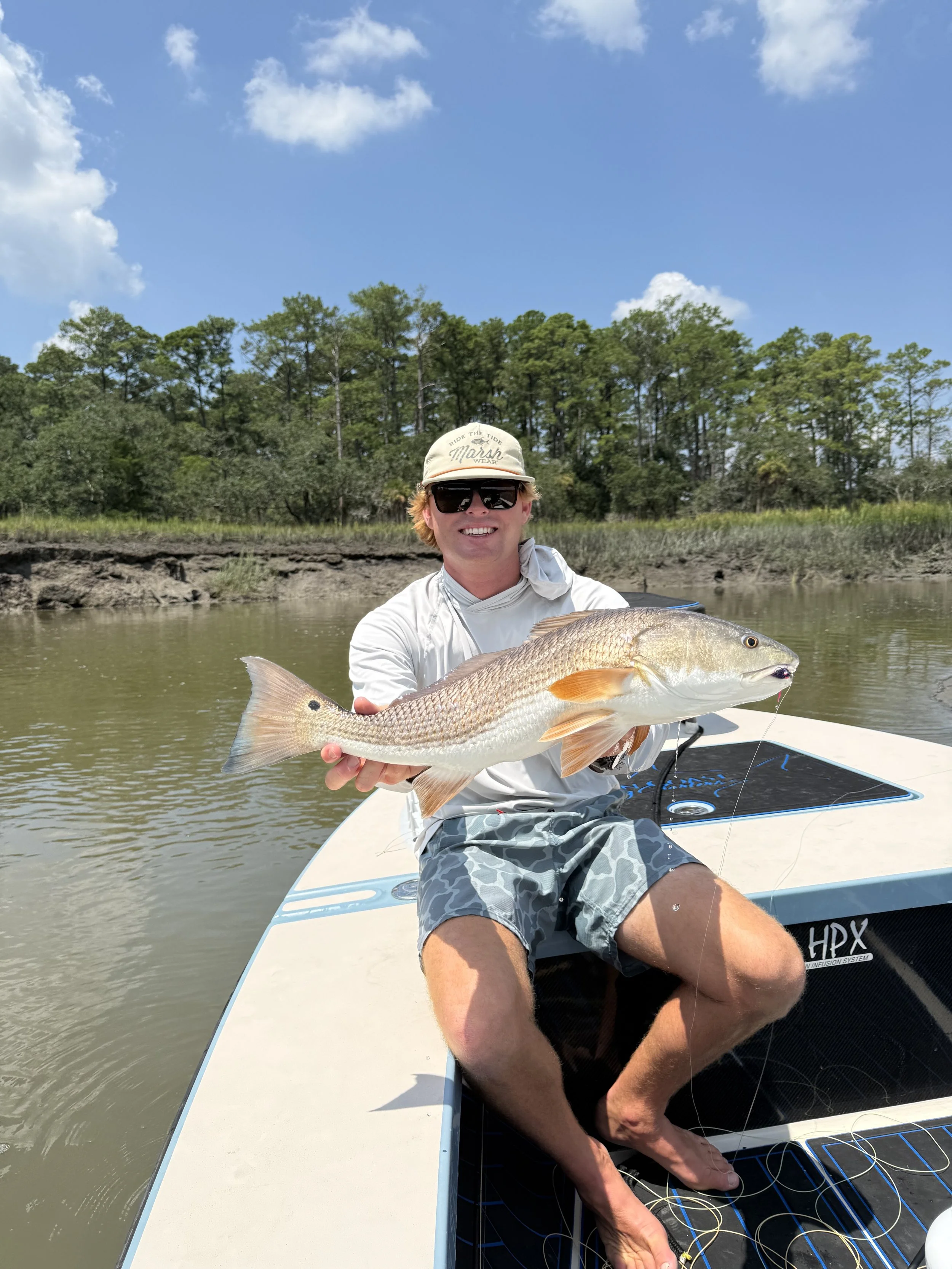 A man in a white shirt, sunglasses, and a beige cap sitting on a boat, proudly holding a large fish he caught, with a body of water and green trees in the background on a sunny day.