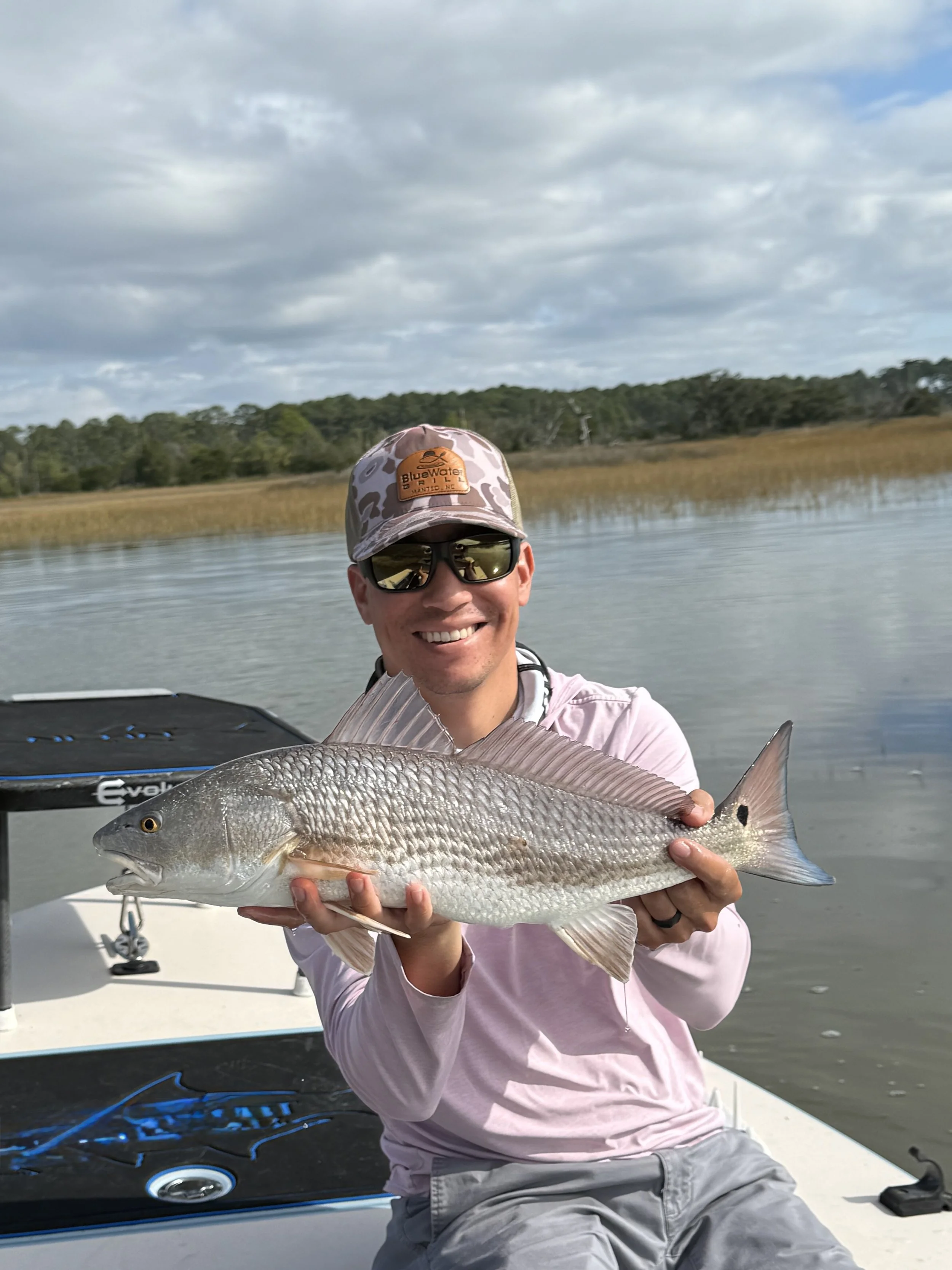 A man holding a large fish on a boat with water and marshland in the background, under a cloudy sky.