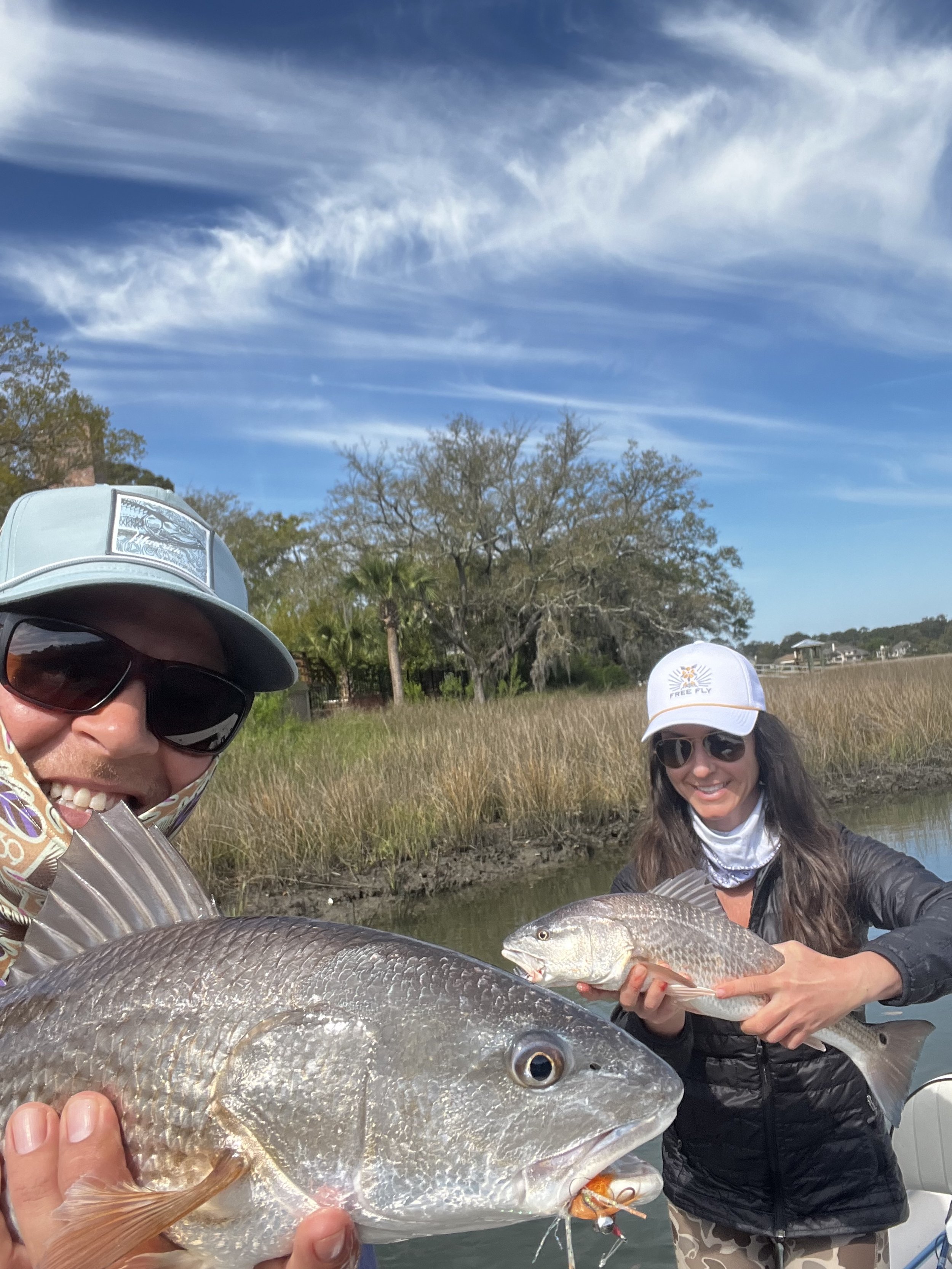 Two women outdoors holding fish they caught, with water, marsh grass, trees, and sky in the background.