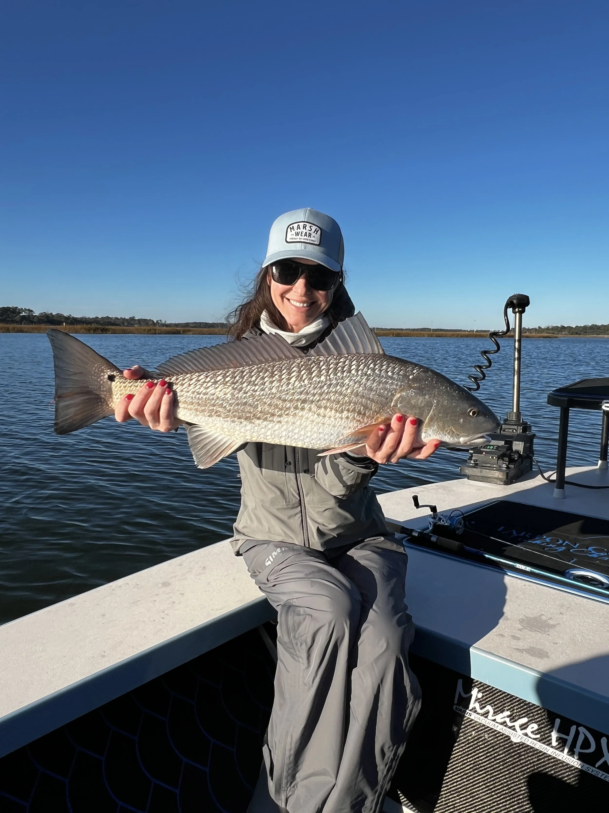 A woman in sunglasses, a cap, and outdoor gear sitting in a boat on a body of water, holding a large fish she caught.