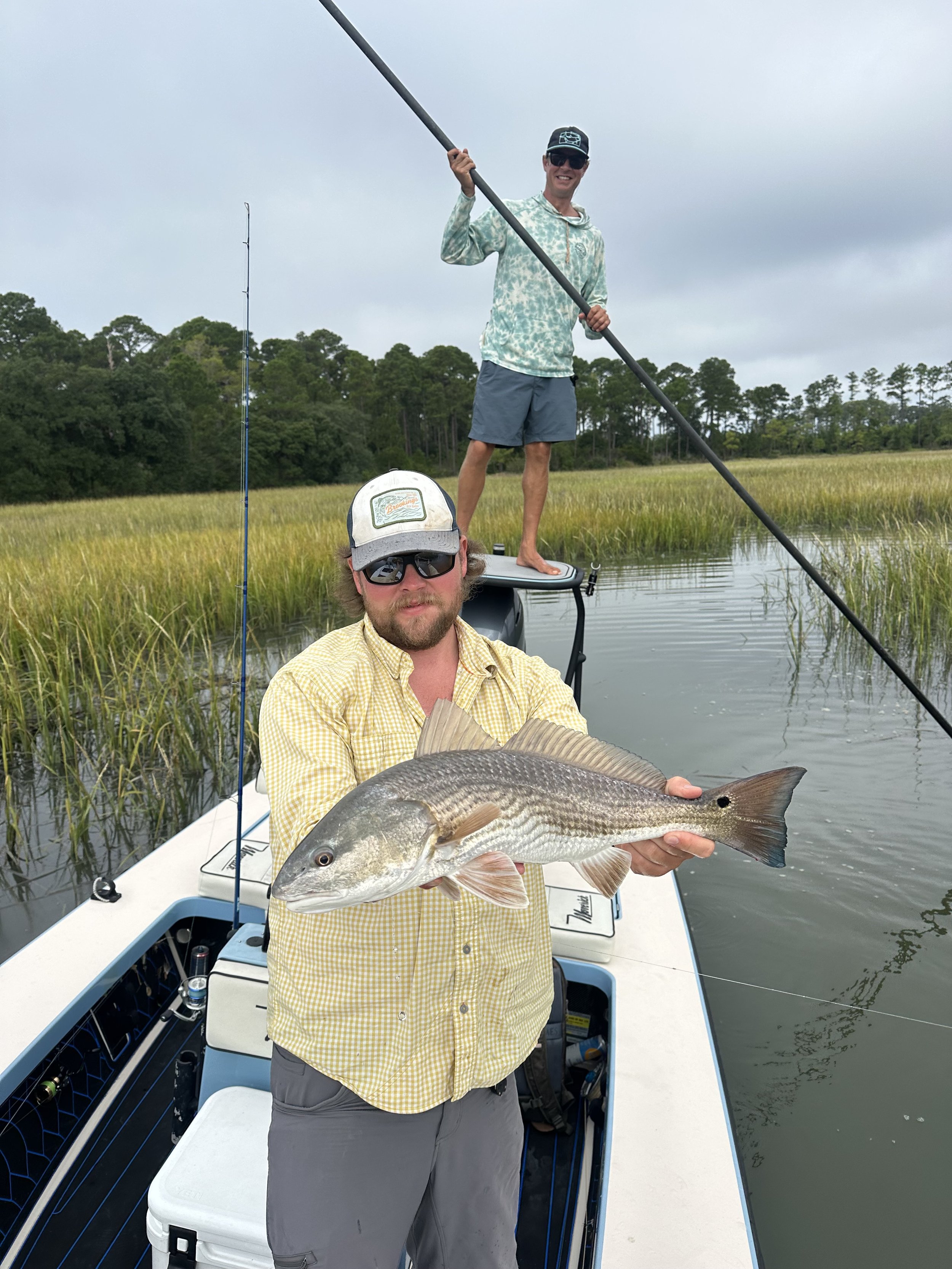A man in a yellow shirt and sunglasses holding a large fish on a boat with another man standing on the boat with a fishing pole and smiling, with marshland and trees in the background.