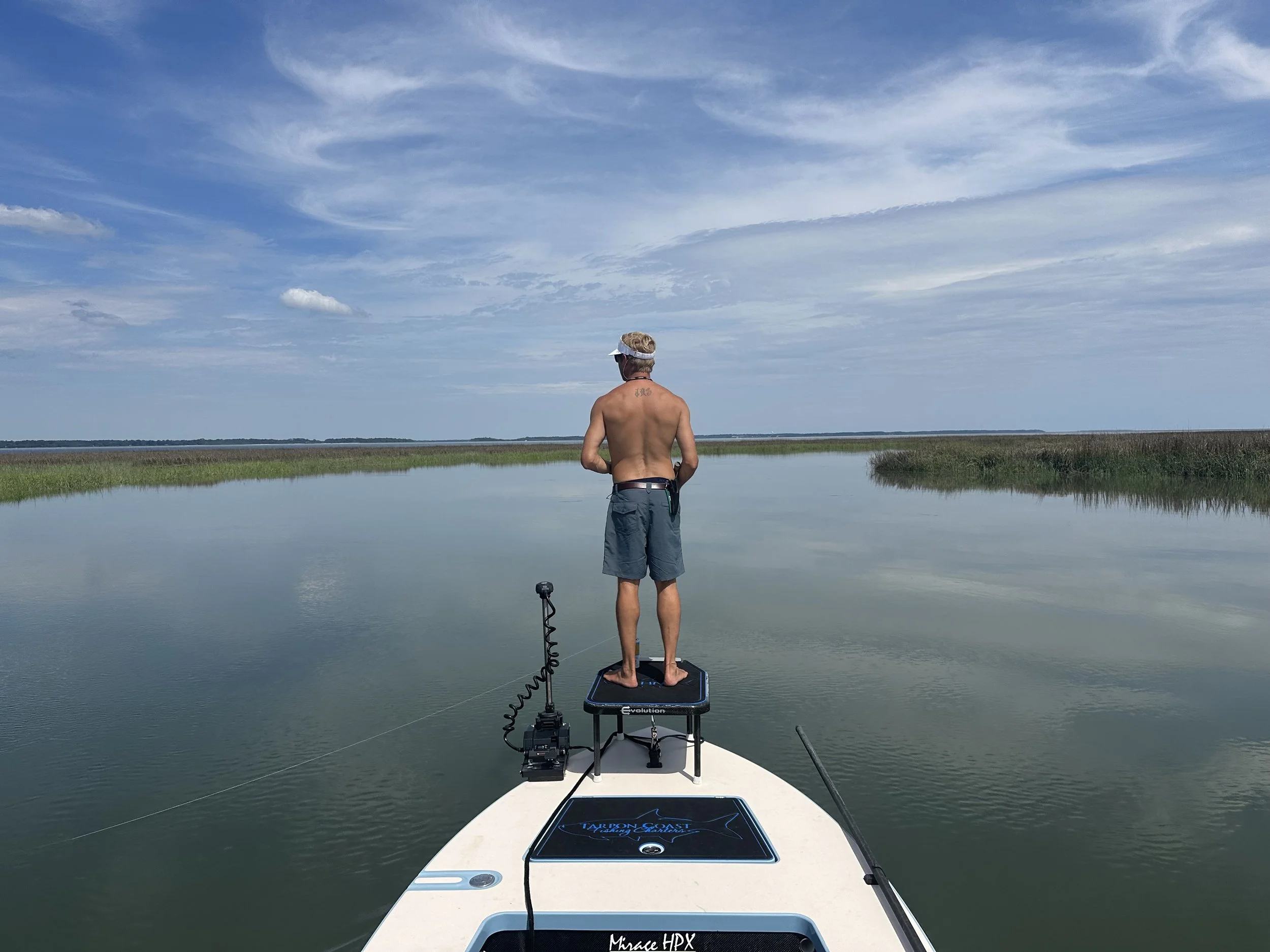 A man standing on a small paddleboard or fishing platform on a calm body of water, facing away, with marshland on either side and a partly cloudy sky above.
