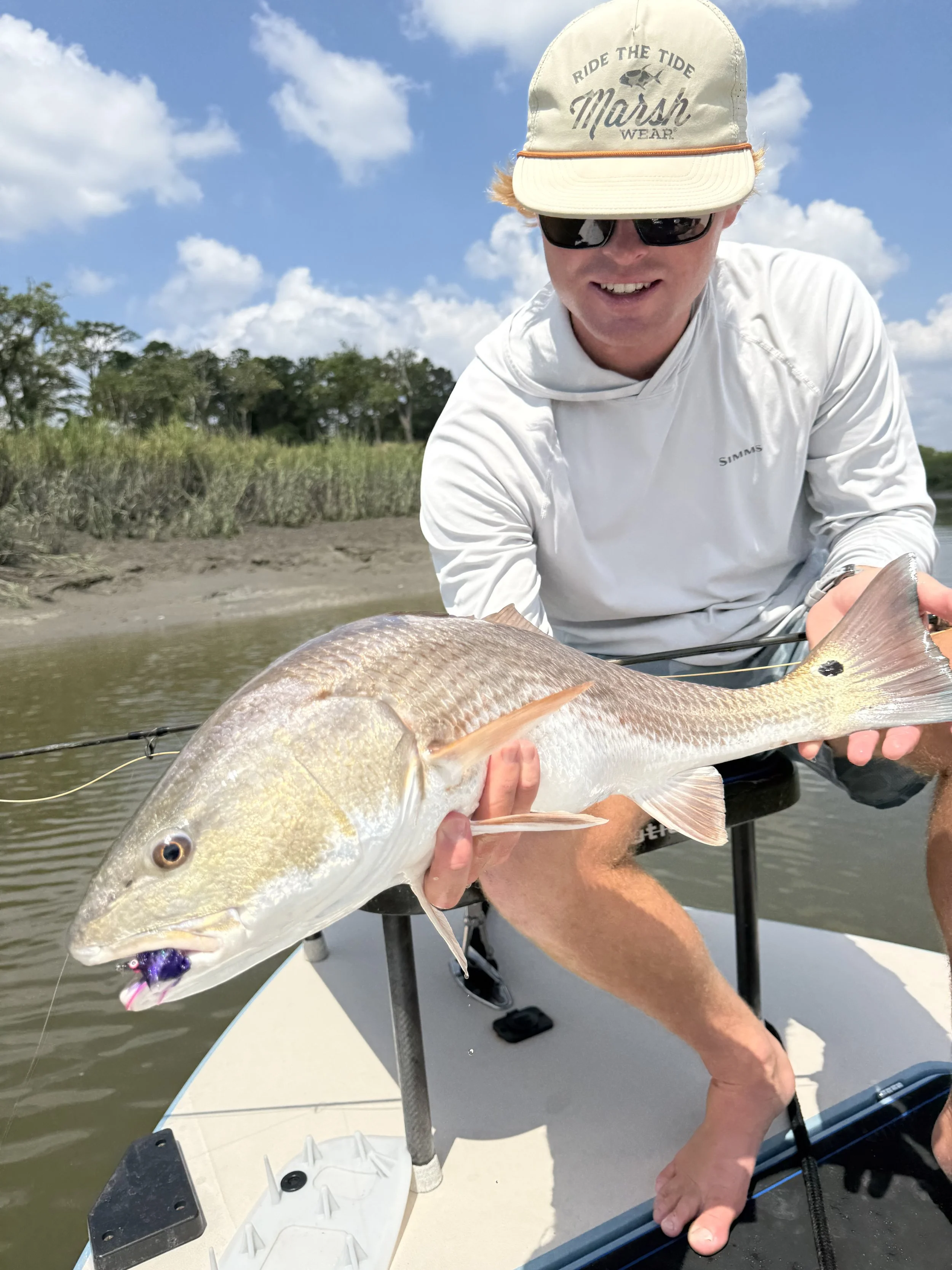 Man holding a large fish on a boat with water and trees in the background on a sunny day.