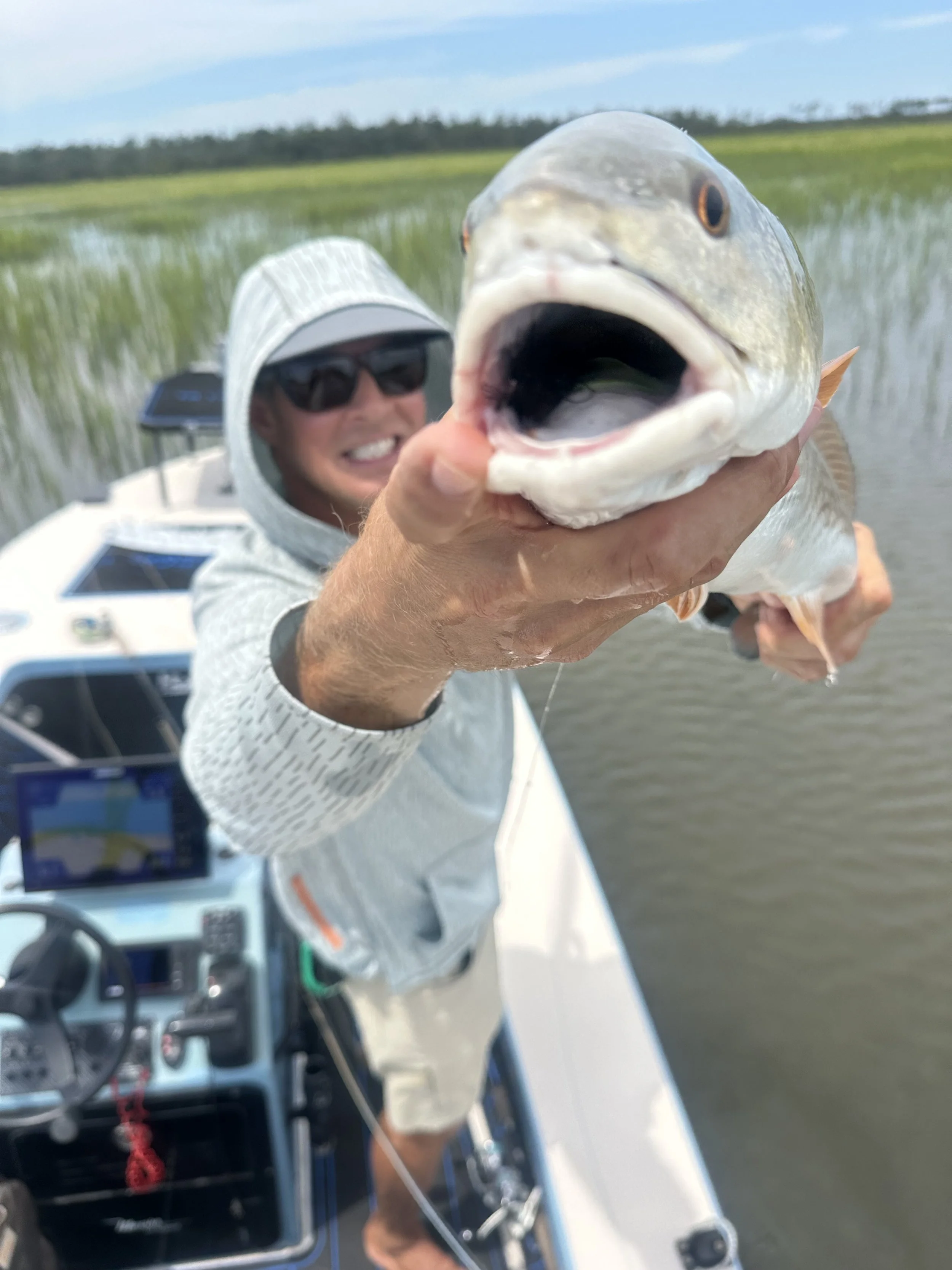Man holding a large fish on a boat in a marsh, smiling, wearing sunglasses and a white hoodie