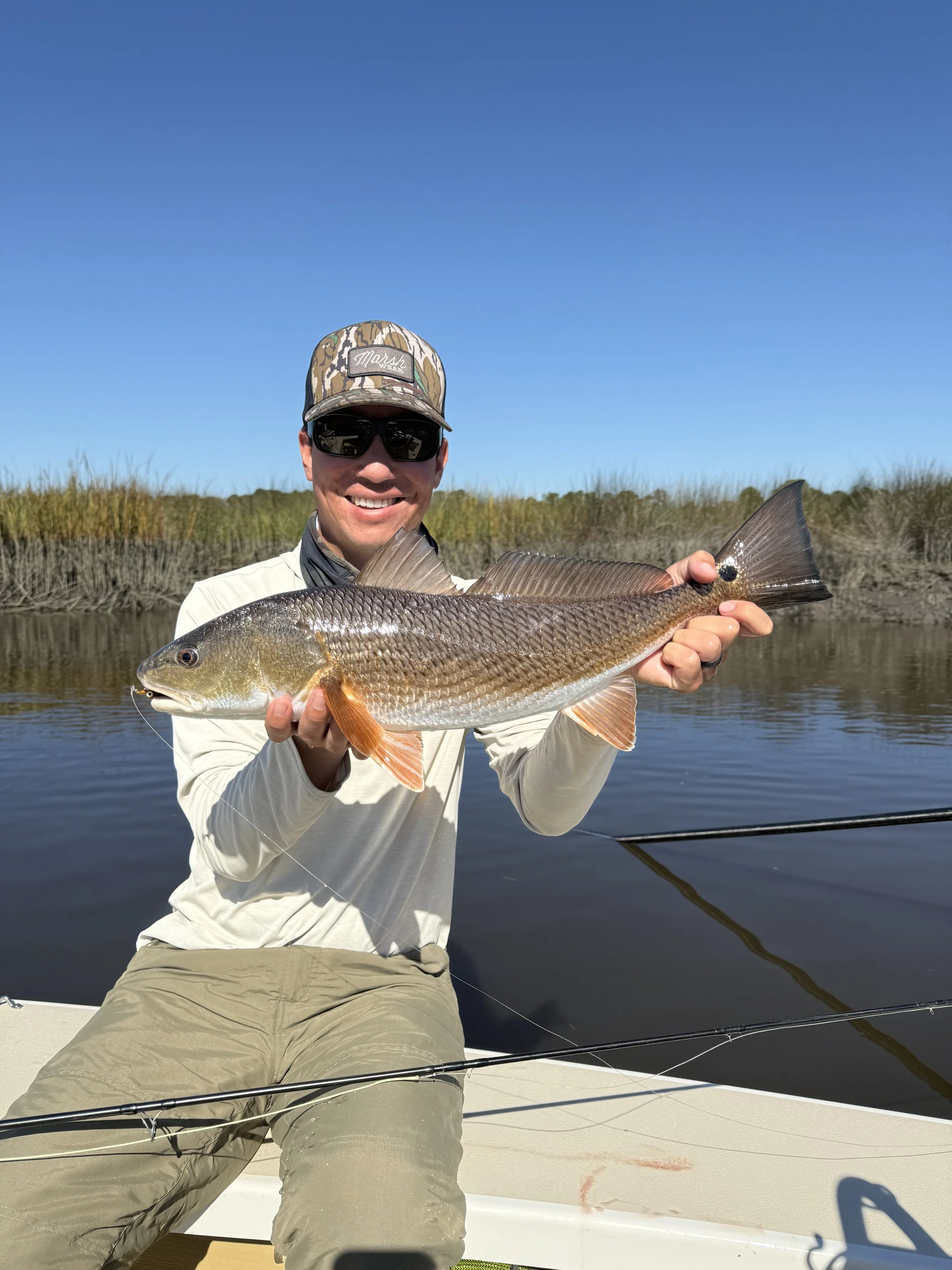 Man in sunglasses and camouflage cap holding a large fish on a boat with water and marsh grass in the background.