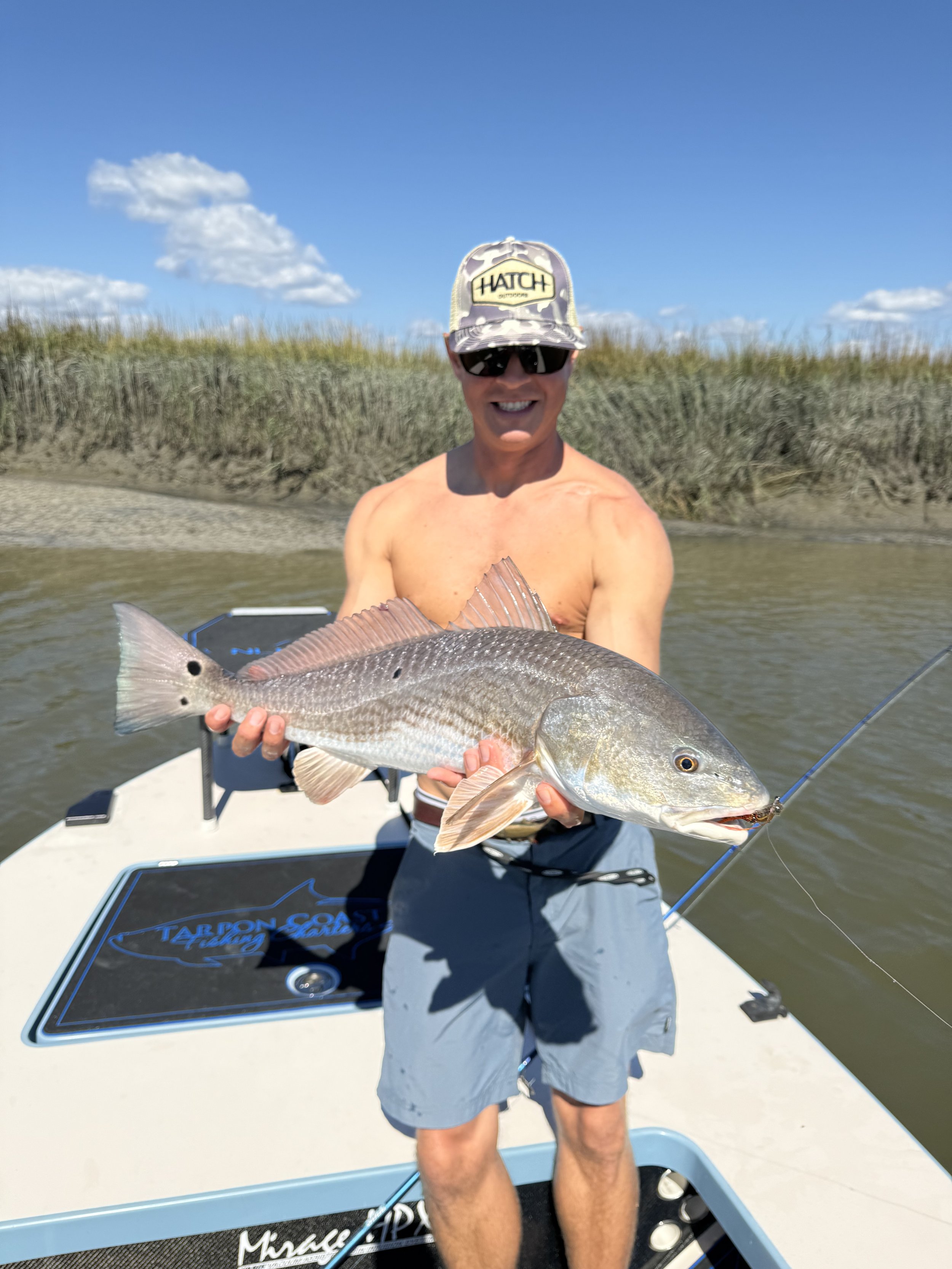 Smiling man in camouflage hat and sunglasses on boat holding a large fish with a sandy shoreline and grassy dunes in background.