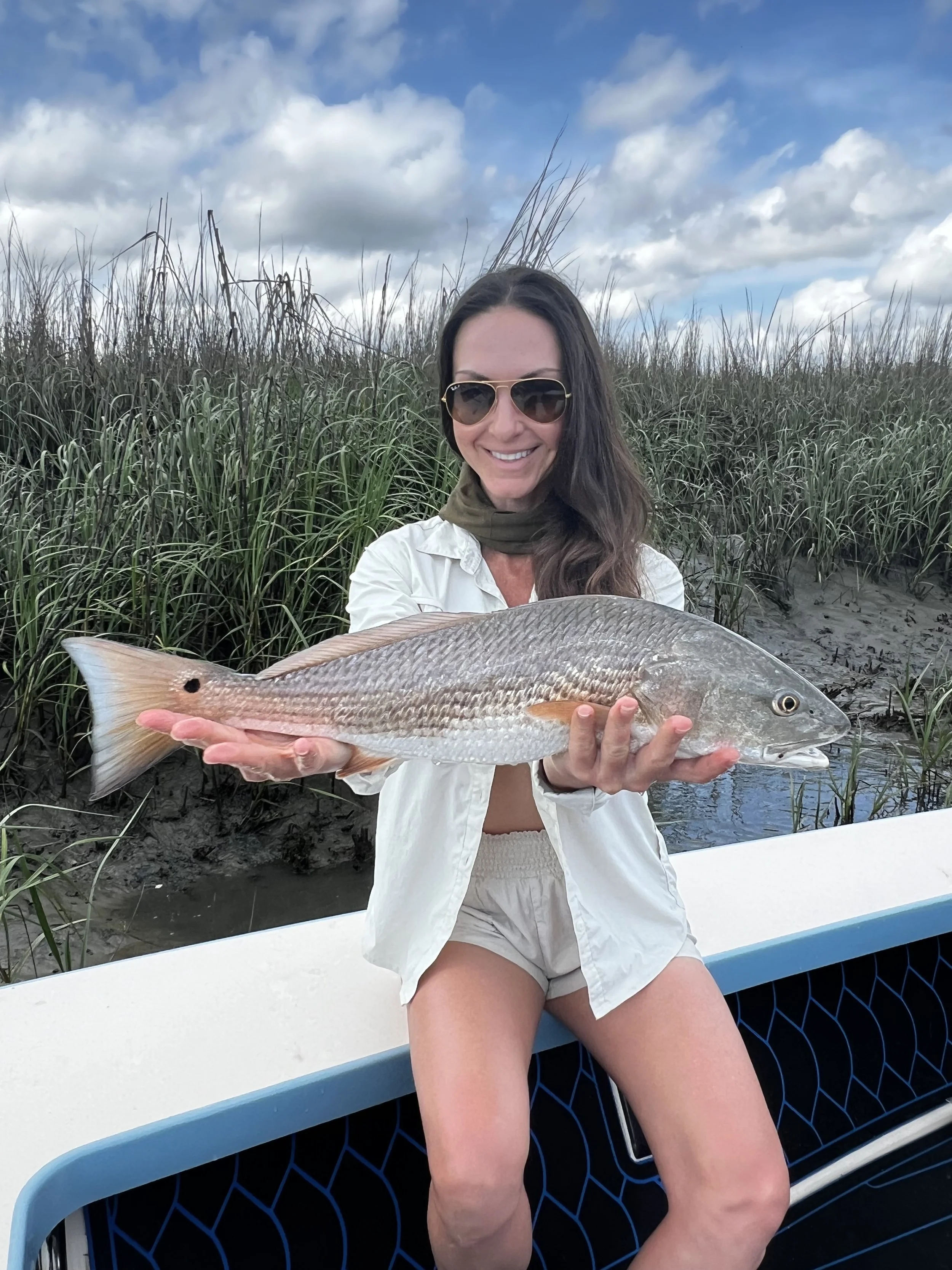 Woman wearing sunglasses smiling and holding a large fish on a boat with marshy grass and cloudy sky in the background.