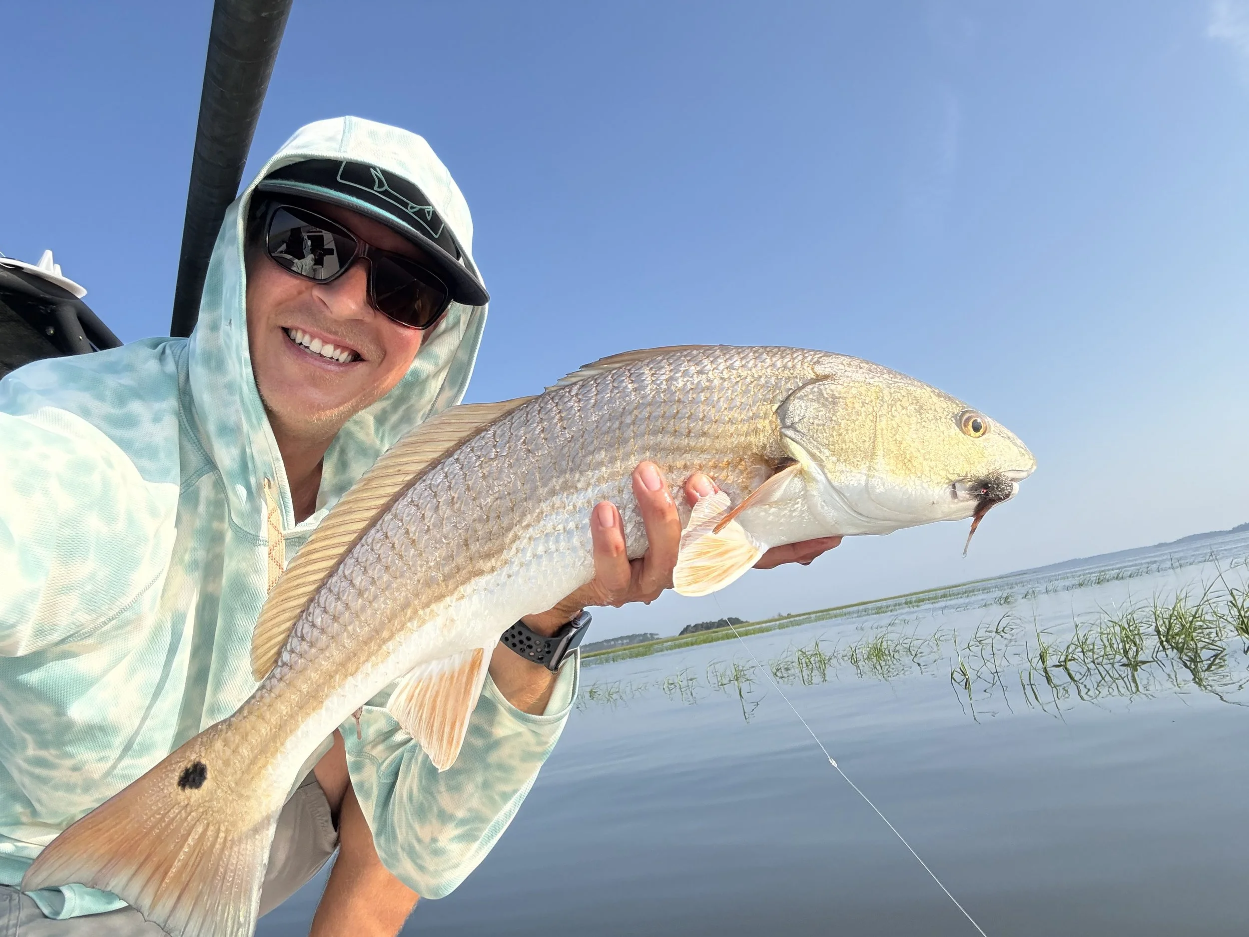 Smiling man in light-colored hoodie and sunglasses holding a large fish near a body of water with a clear sky