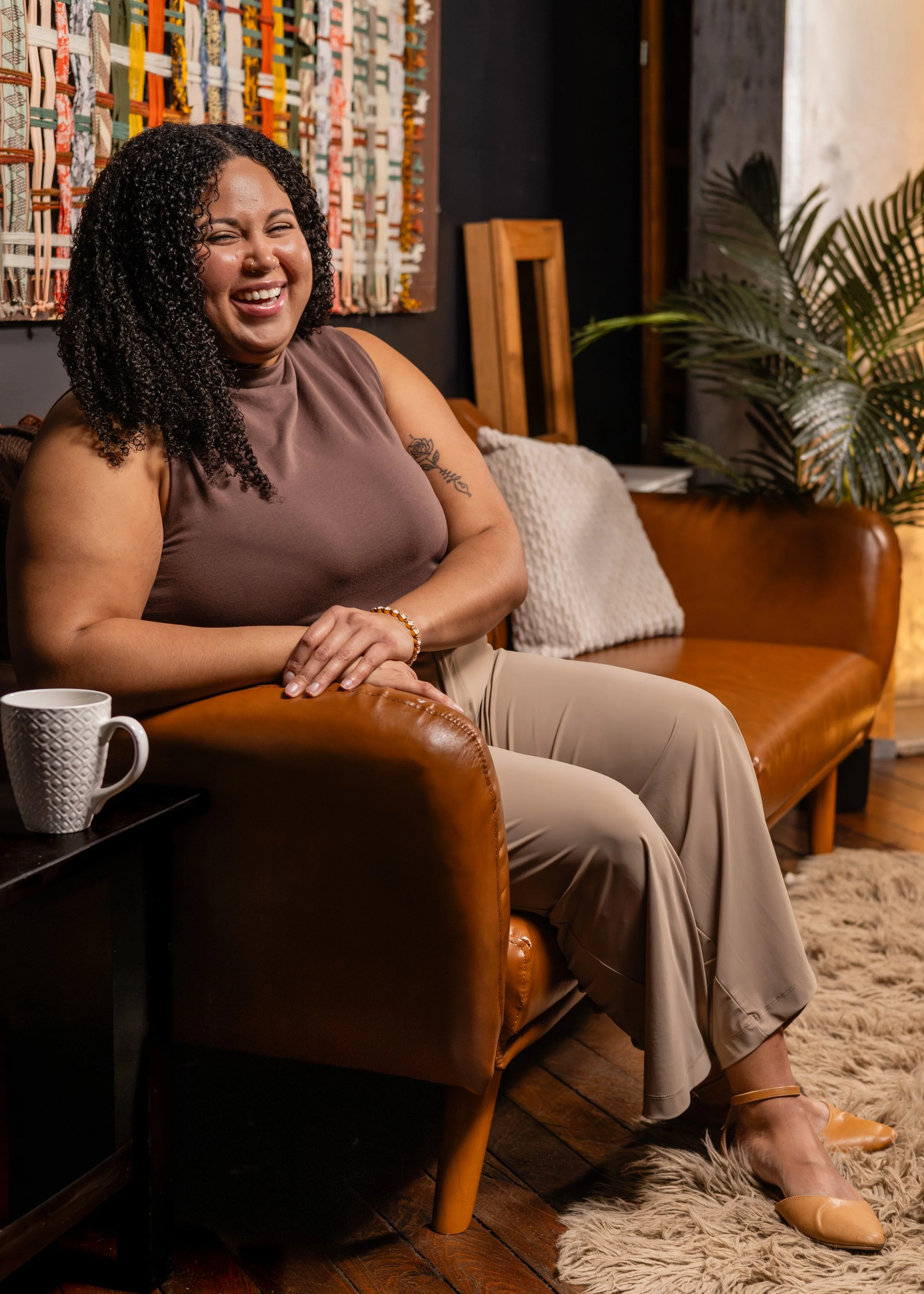 A woman with curly hair, sitting on a leather couch, smiling and looking at the camera. There is a white mug on a small table beside her, and a green plant in the background.