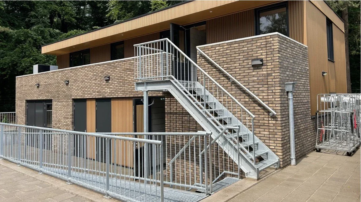 Moderne twee verdieping bijgebouw met trap, houten en bakstenen gevels, metalen trap, terrassen, omgeven door bomen.