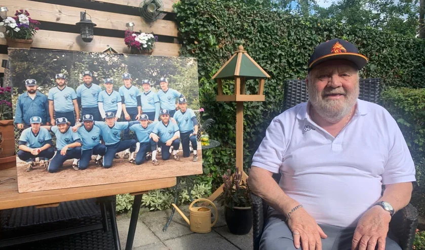 Een man met een witte polo en een baseballpet zit buiten op een stoel, naast een groot teamfoto van een honkbalteam dat op een houtachtig ondergrond staat, omringd door planten en bloemen.