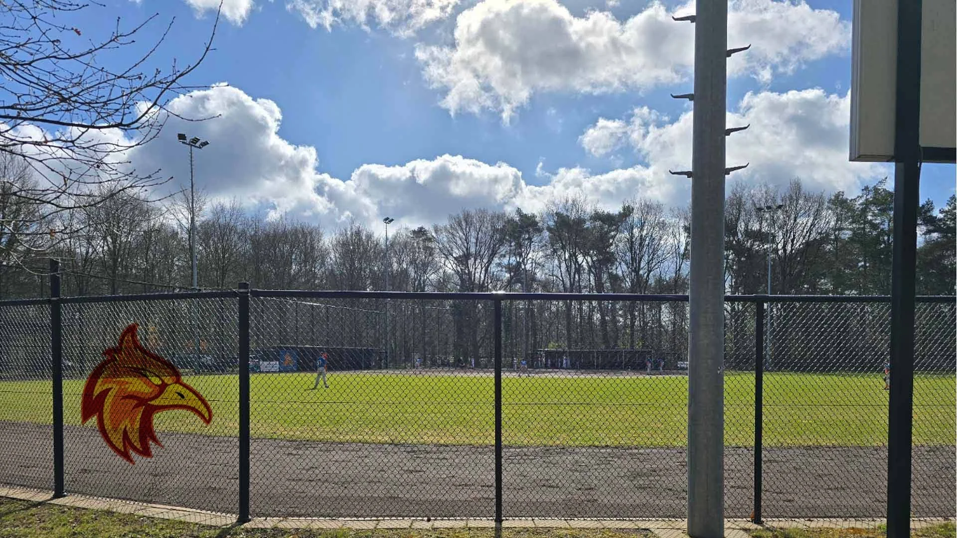 Sportveld met spelers, omgeven door een metalen hek met het firebirds logo, met bomen en een bewolkte hemel op de achtergrond.