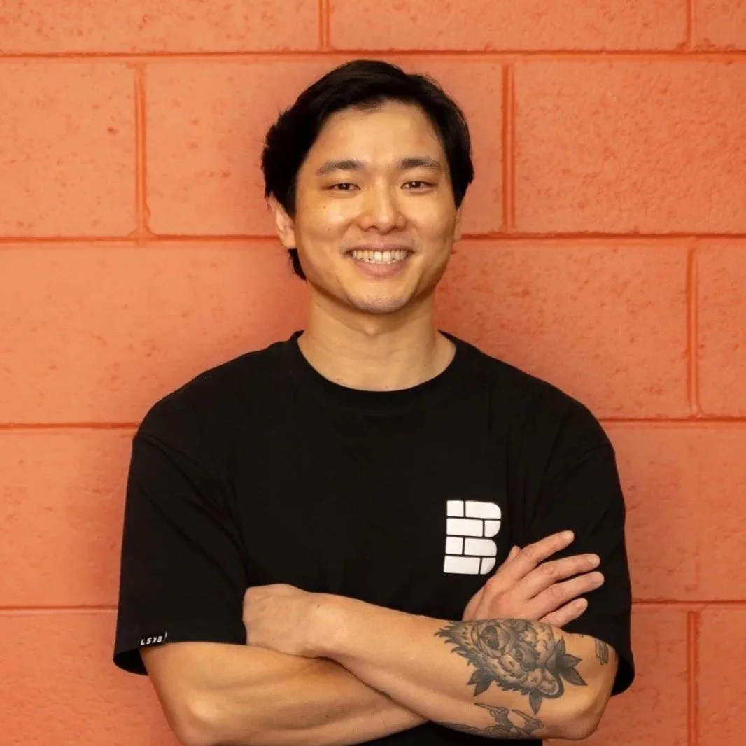 Young man with long hair in a bun, smiling, wearing a black athletic shirt, with a tattoo of a lion's face on his right arm, crossed arms, standing against a dark, blurred background.