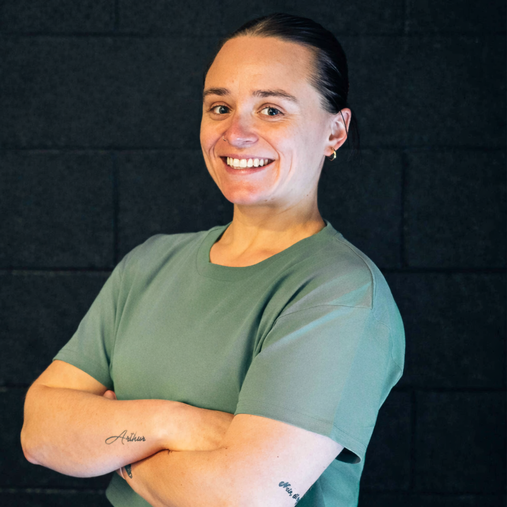 A woman with dark hair pulled back, wearing a green t-shirt and gold hoop earrings, smiling and standing with arms crossed against a dark brick wall background.