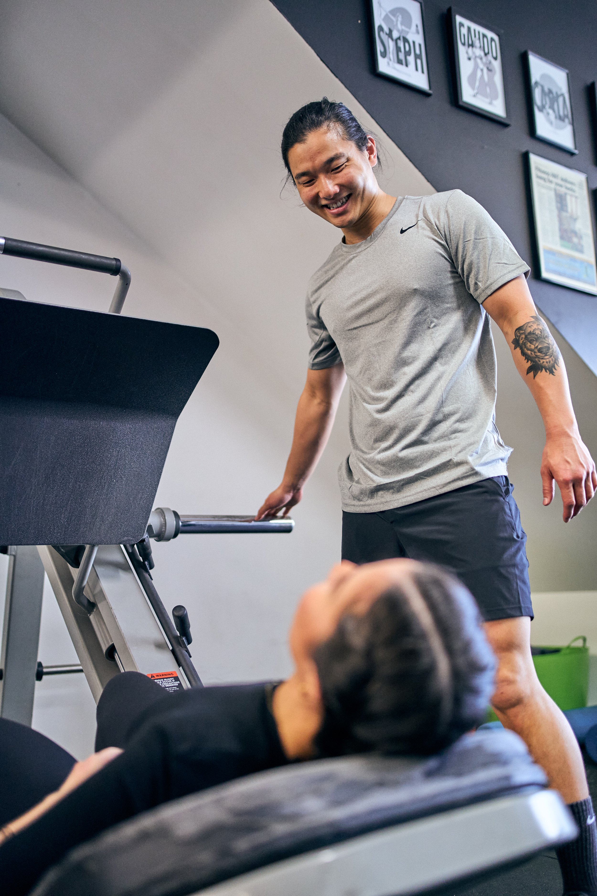 A man with long hair tied back, smiling and leaning on a treadmill, talking with a woman lying on a bench in a gym.