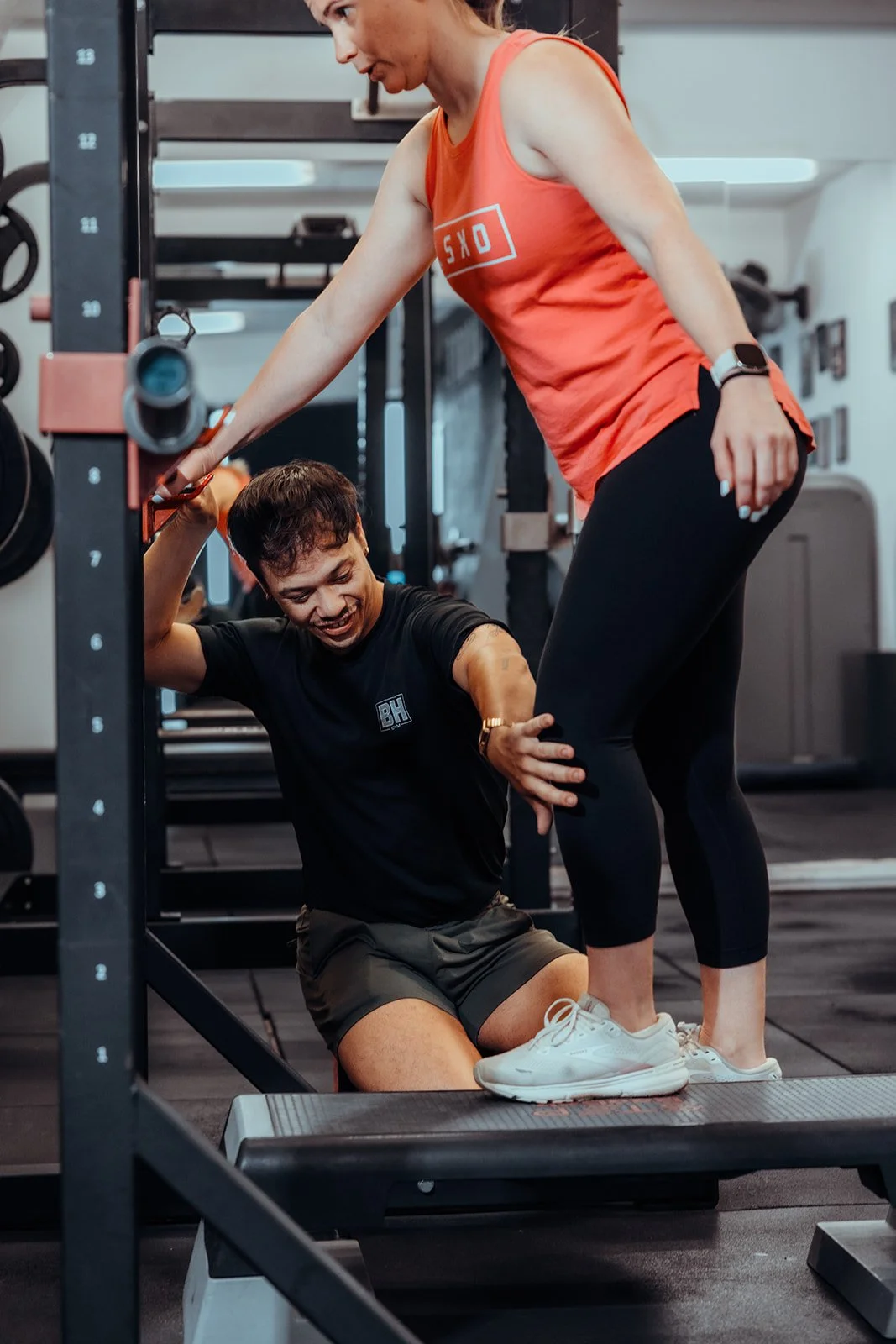 A woman assisting a man during a workout in a gym. The woman is holding the man's arm for support as he performs a exercise on a step platform. Both are smiling and focused on the activity.