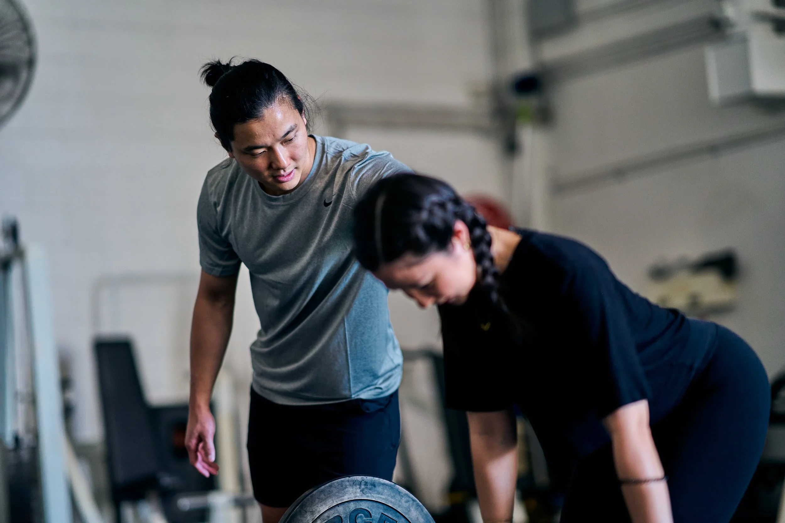 A personal trainer instructs a woman during her workout in a gym, with the woman lifting a barbell.