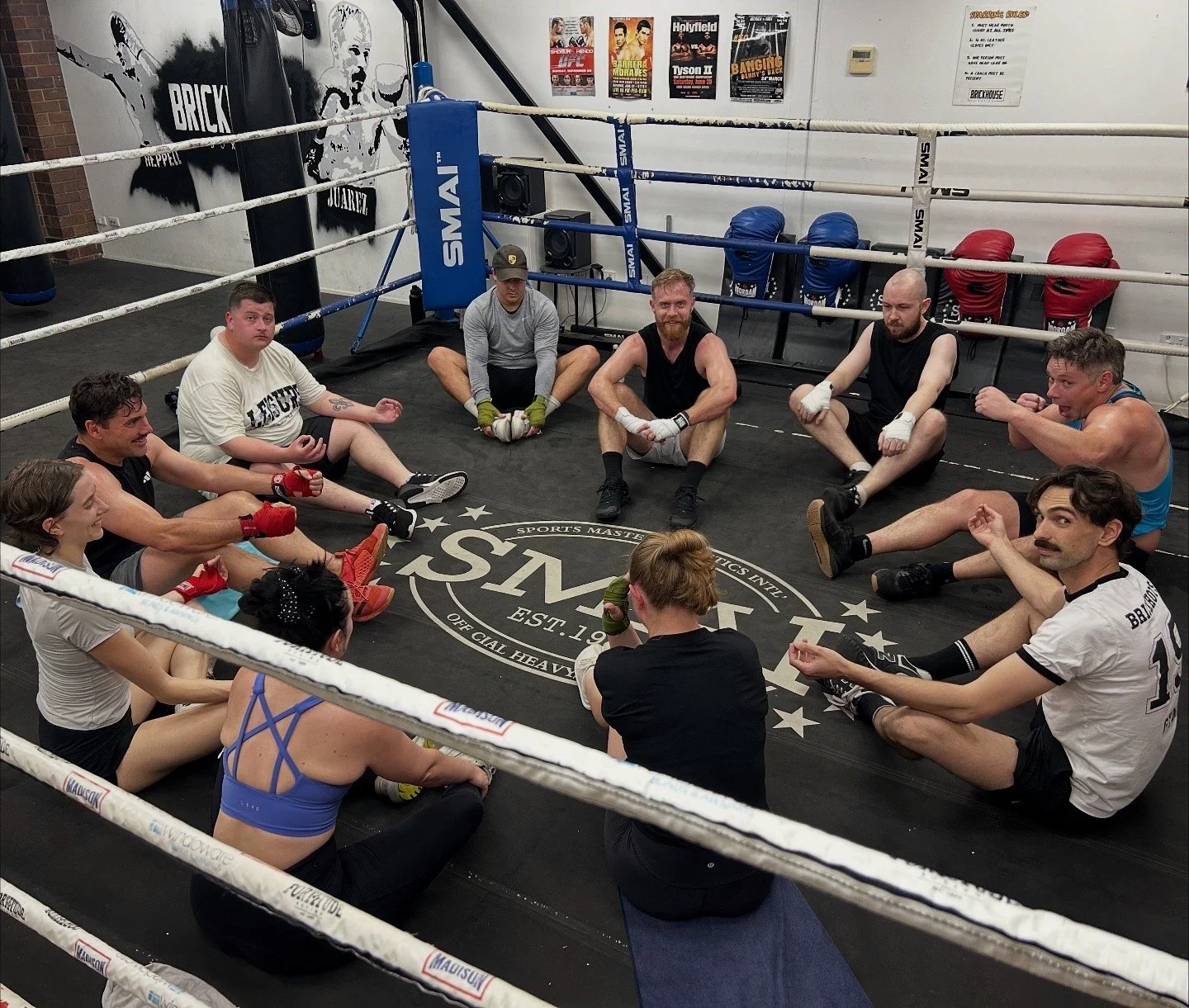 A group of people sitting in a circle on a boxing gym floor, participating in a discussion or training session. The gym has a boxing ring with ropes and punching bags in the background.
