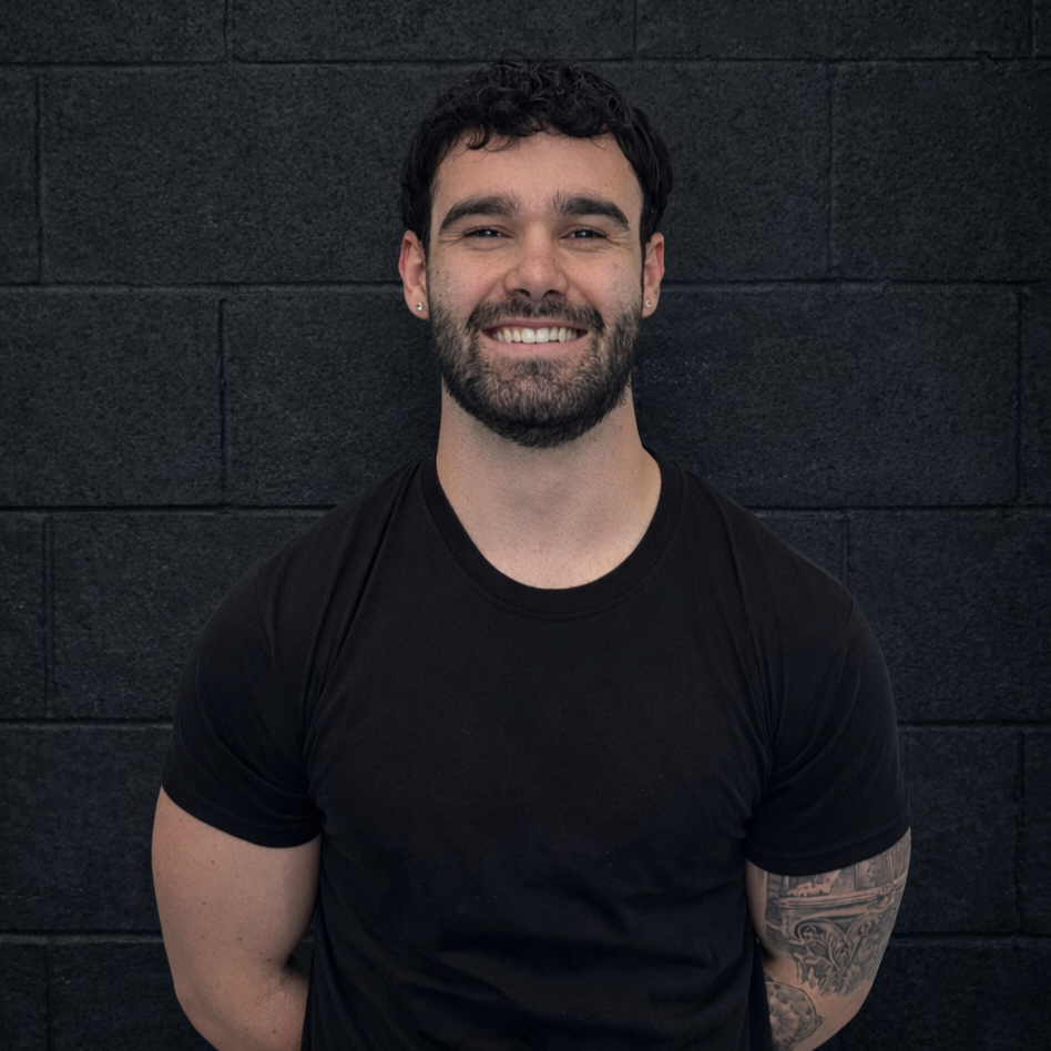 A smiling man with dark curly hair and a beard, wearing a black t-shirt, standing in front of a dark brick wall.