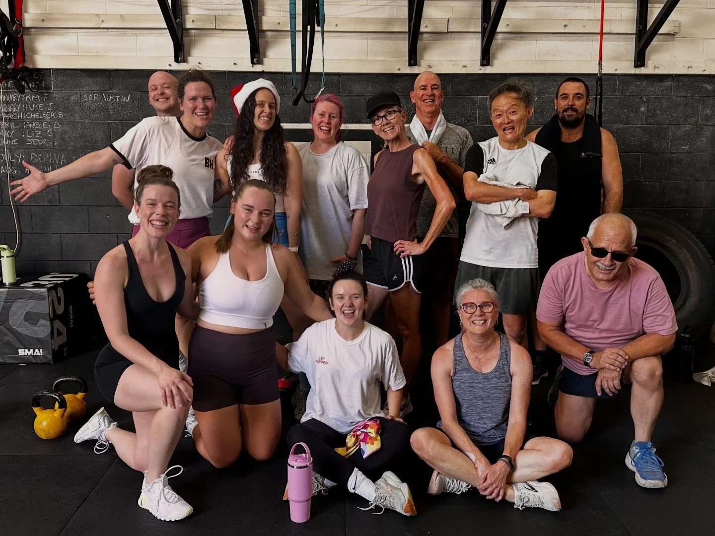 Group of people in workout attire posing inside a gym, smiling, some wearing glasses, with kettlebells and workout equipment in the background.