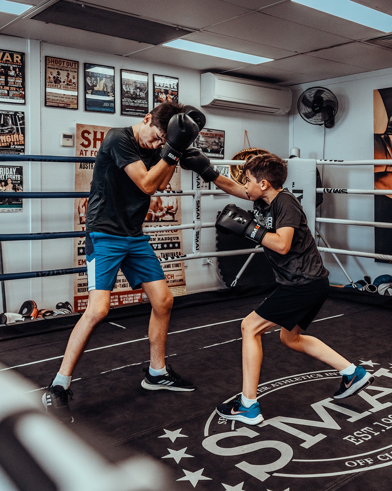 A young boy and a young man are boxing in a gym ring. The boy is throwing a punch at the young man, who is blocking it with his boxing gloves. The gym has posters on the wall and boxing equipment on the floor.
