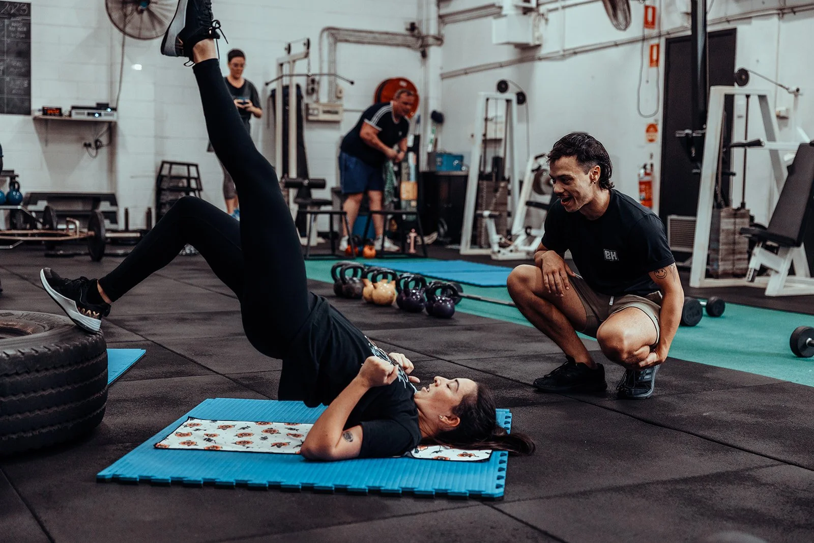 A woman lying on her back on a blue exercise mat, lifting her hips and legs in a workout, while a trainer kneels beside her, encouraging her. Other people are working out in the background in a gym.