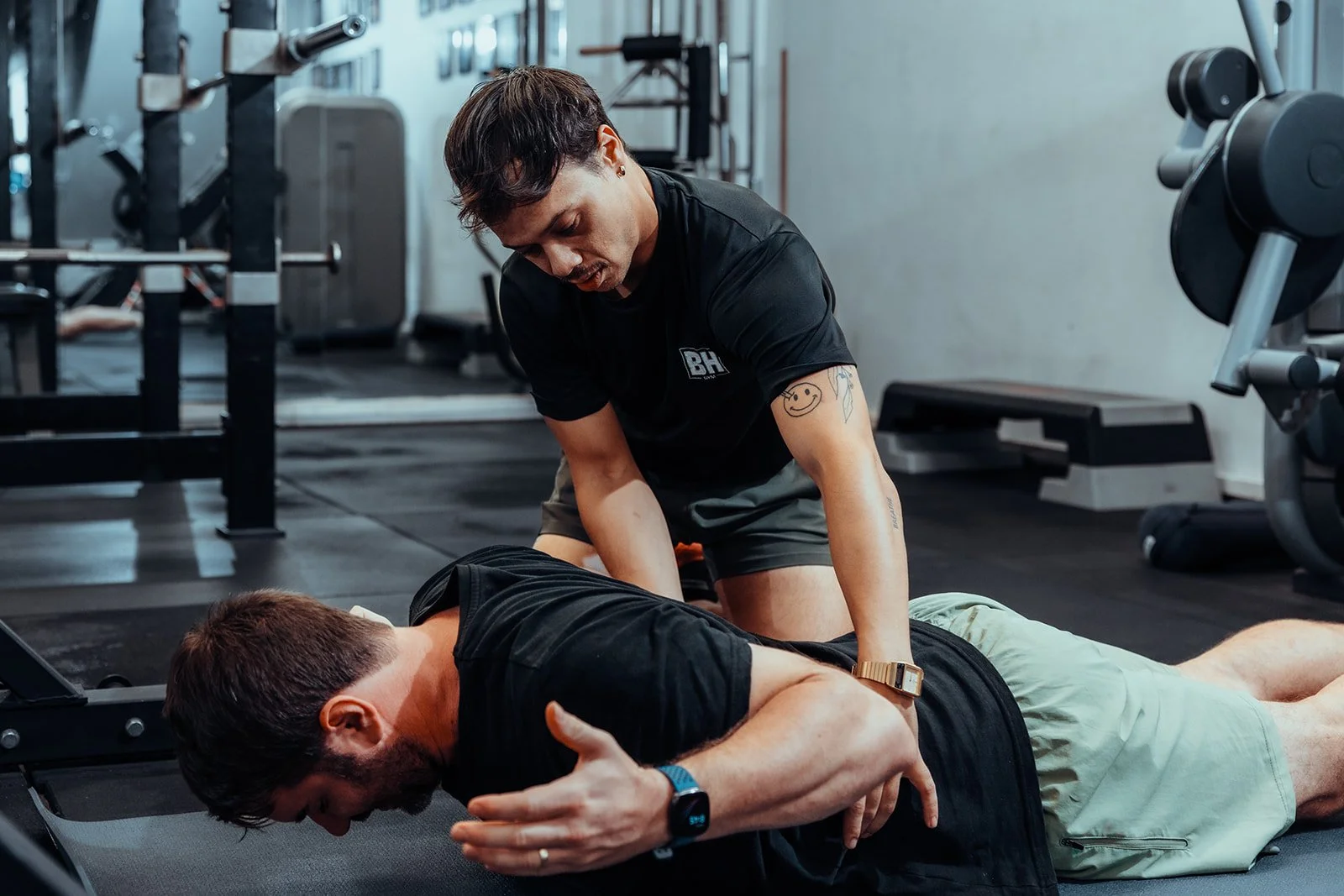 A personal trainer assisting a man with a sit-up in a gym, with gym equipment visible in the background.