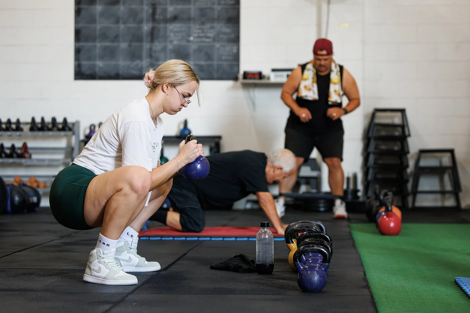 Woman in workout attire squatting and lifting a kettlebell while two men exercising in the background in a gym.