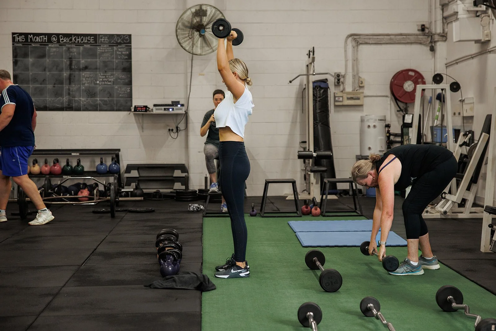 Women working out in a gym, lifting dumbbells and using equipment on a green mat, with other people exercising in the background.