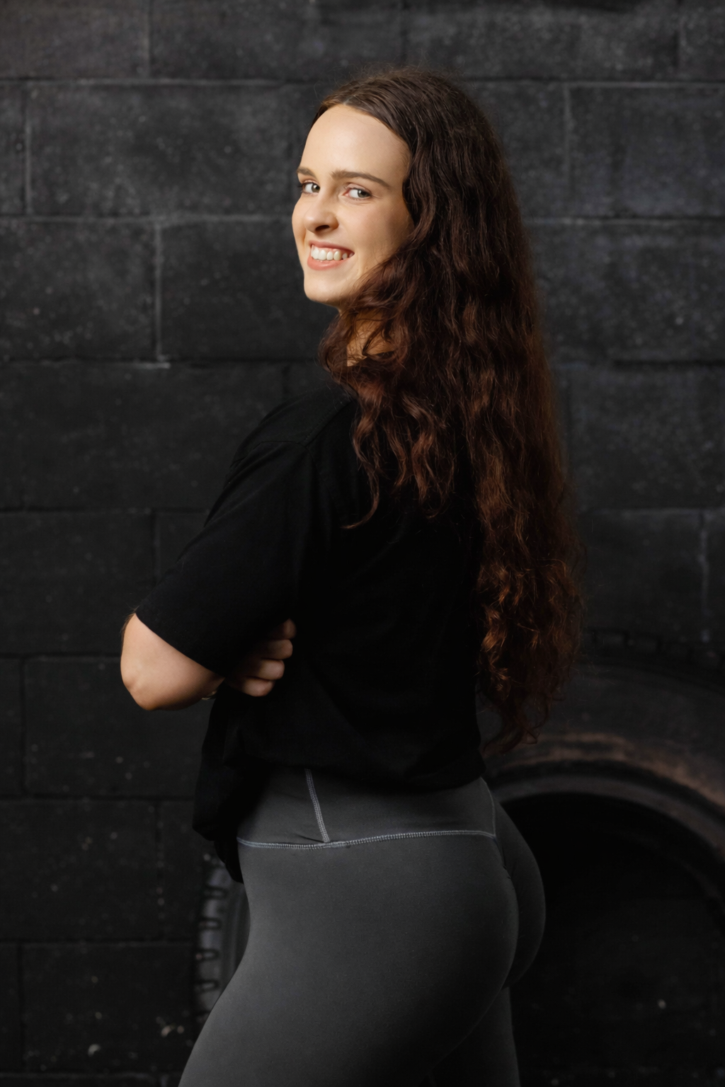 A woman with long, wavy brown hair, wearing a black t-shirt and gray leggings, standing against a dark brick wall, smiling and turning slightly to the side.