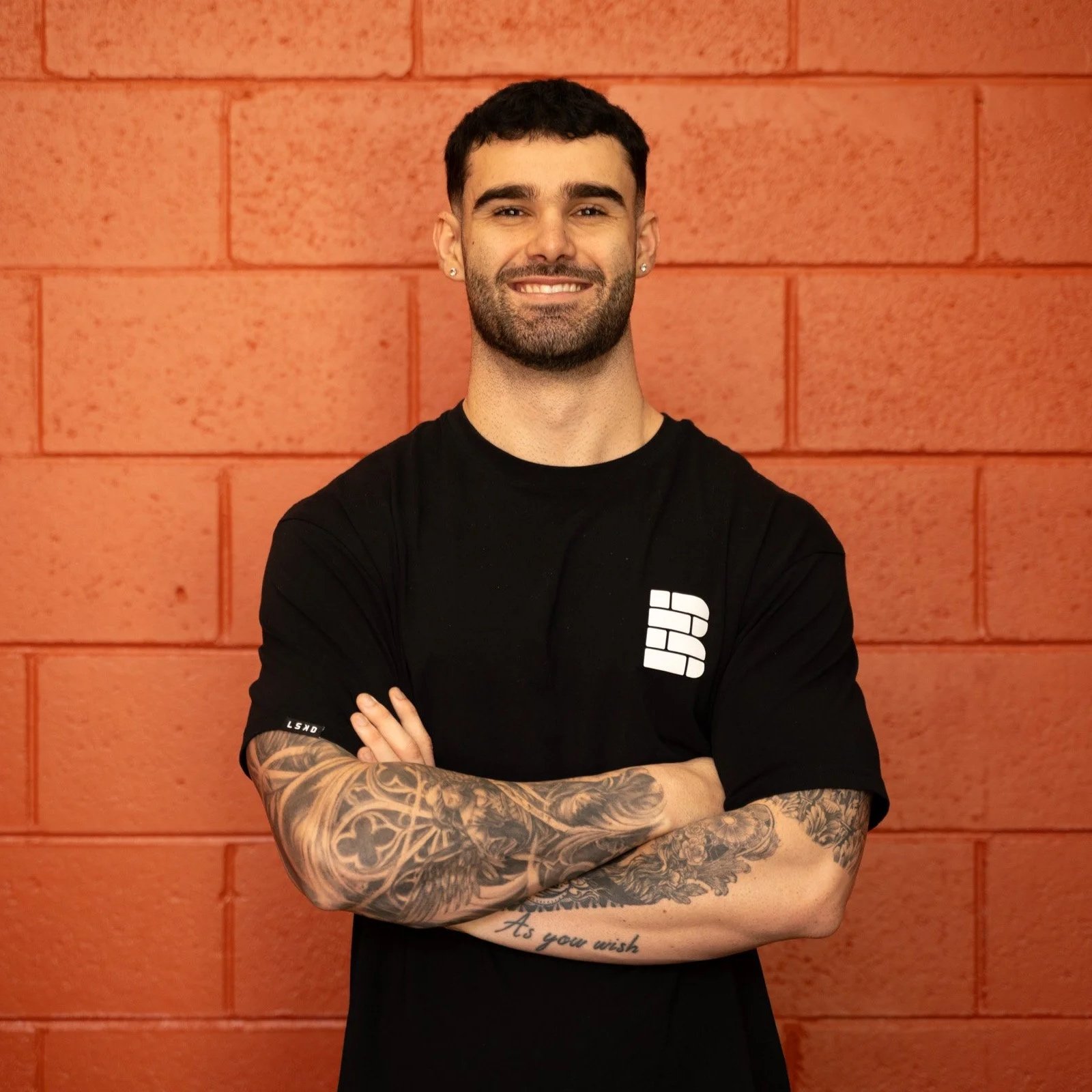 A smiling man with dark curly hair and a beard, wearing a black t-shirt, standing in front of a dark brick wall.