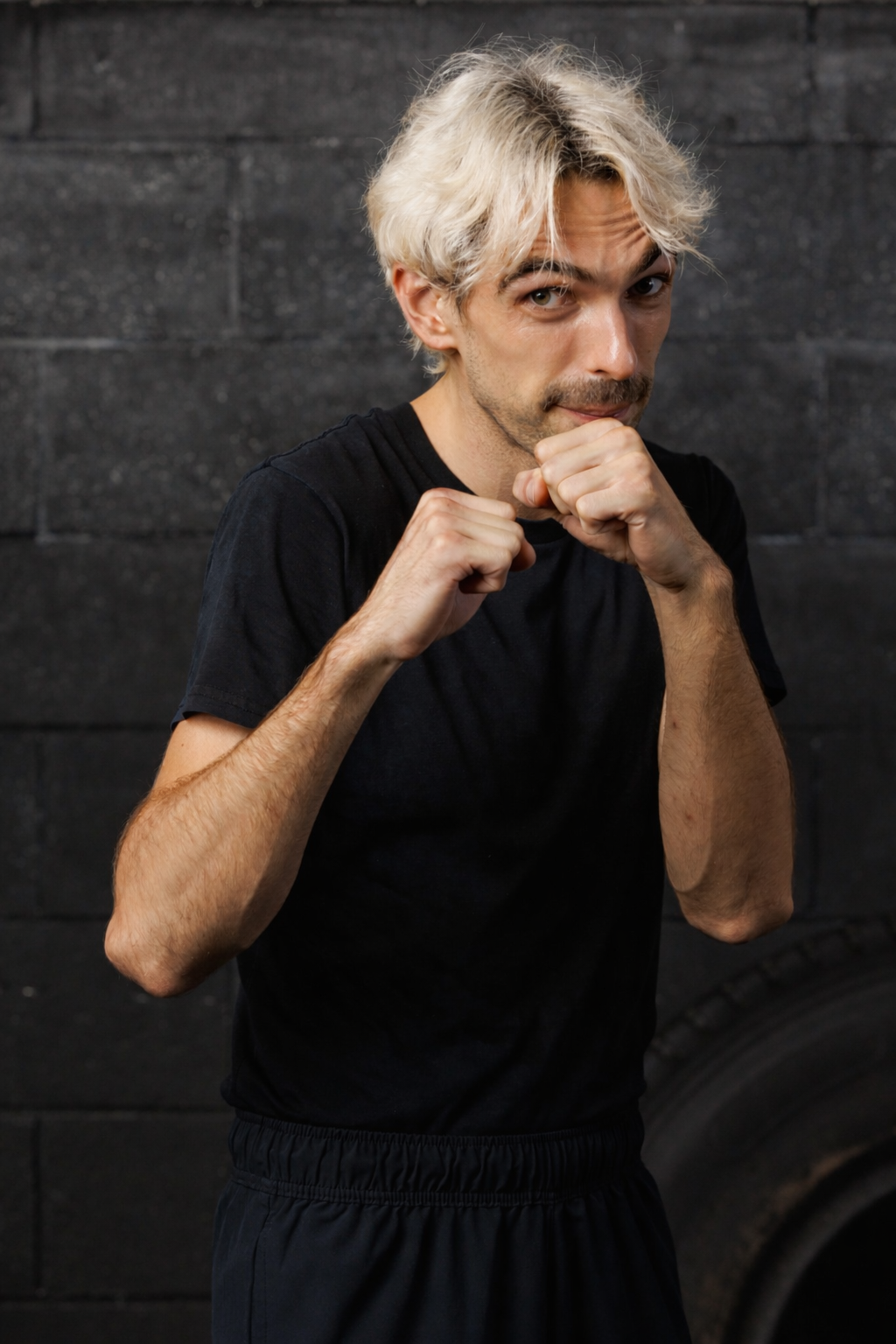 A young man with blond hair and a black t-shirt in a boxing stance, with fists raised, in front of a dark brick wall.