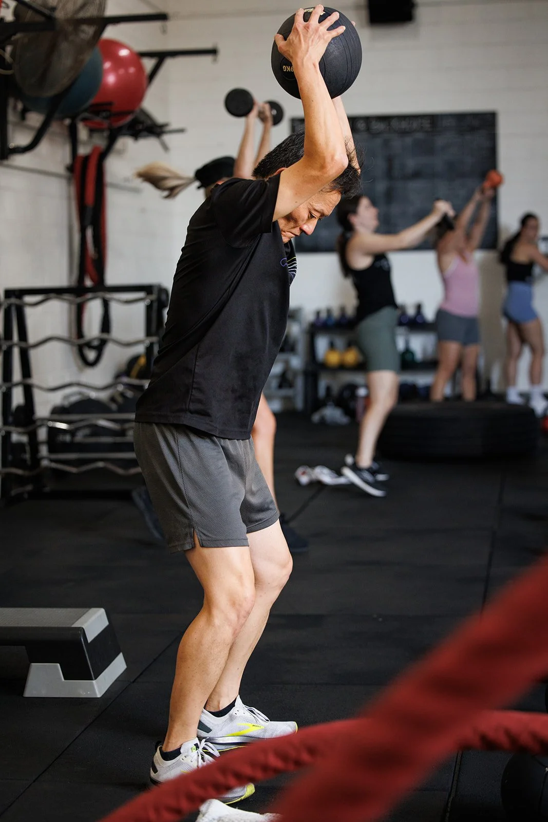 Woman in black t-shirt and gray shorts doing a medicine ball overhead in a gym.