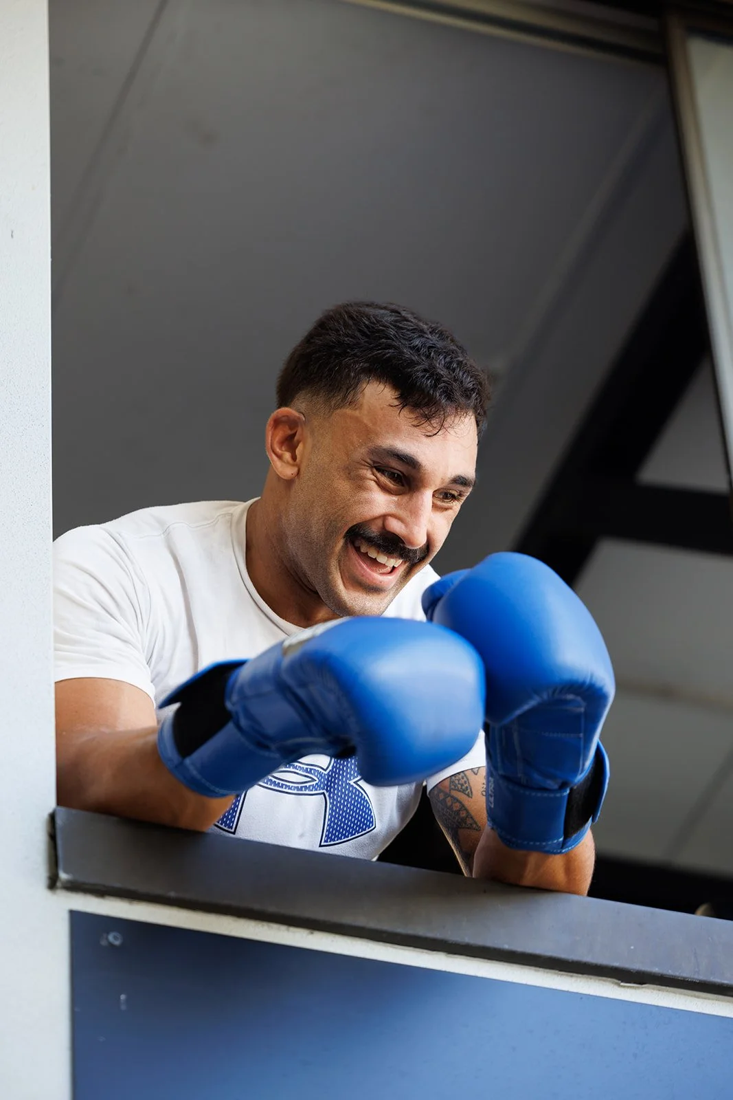 A man with dark hair and a mustache, wearing blue boxing gloves, smiling and leaning on a railing.