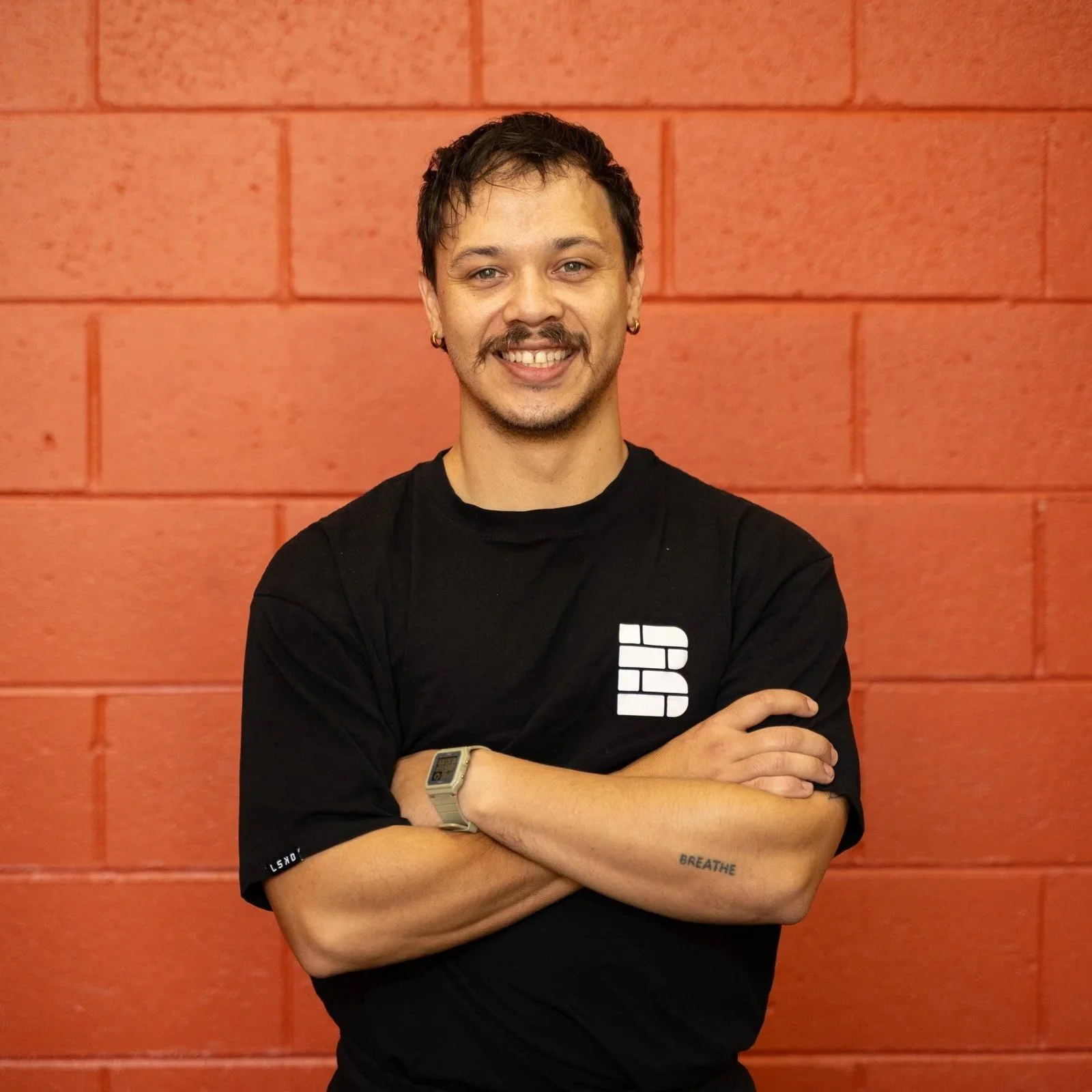 A young man with short dark hair, wearing a black T-shirt, gold watch, and olive green pants, standing with arms crossed in front of a black wall and tires, smiling at the camera.