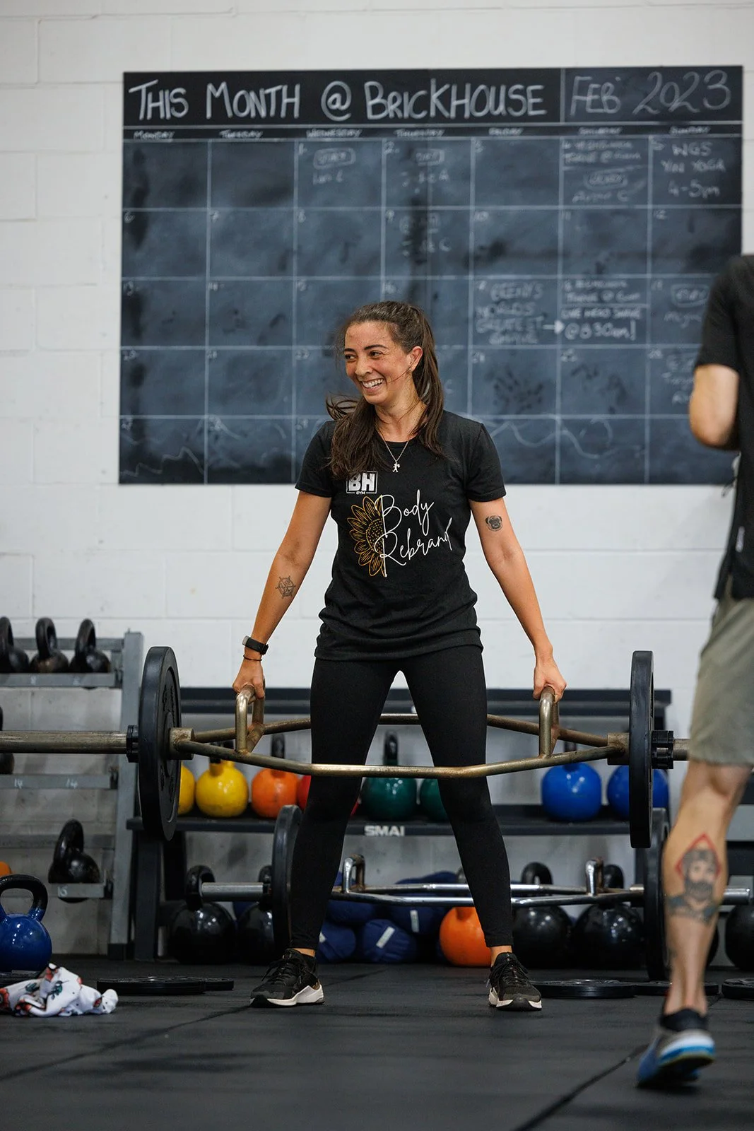 Woman smiling while holding a barbell in a gym with kettlebells on racks and a blackboard calendar in the background.