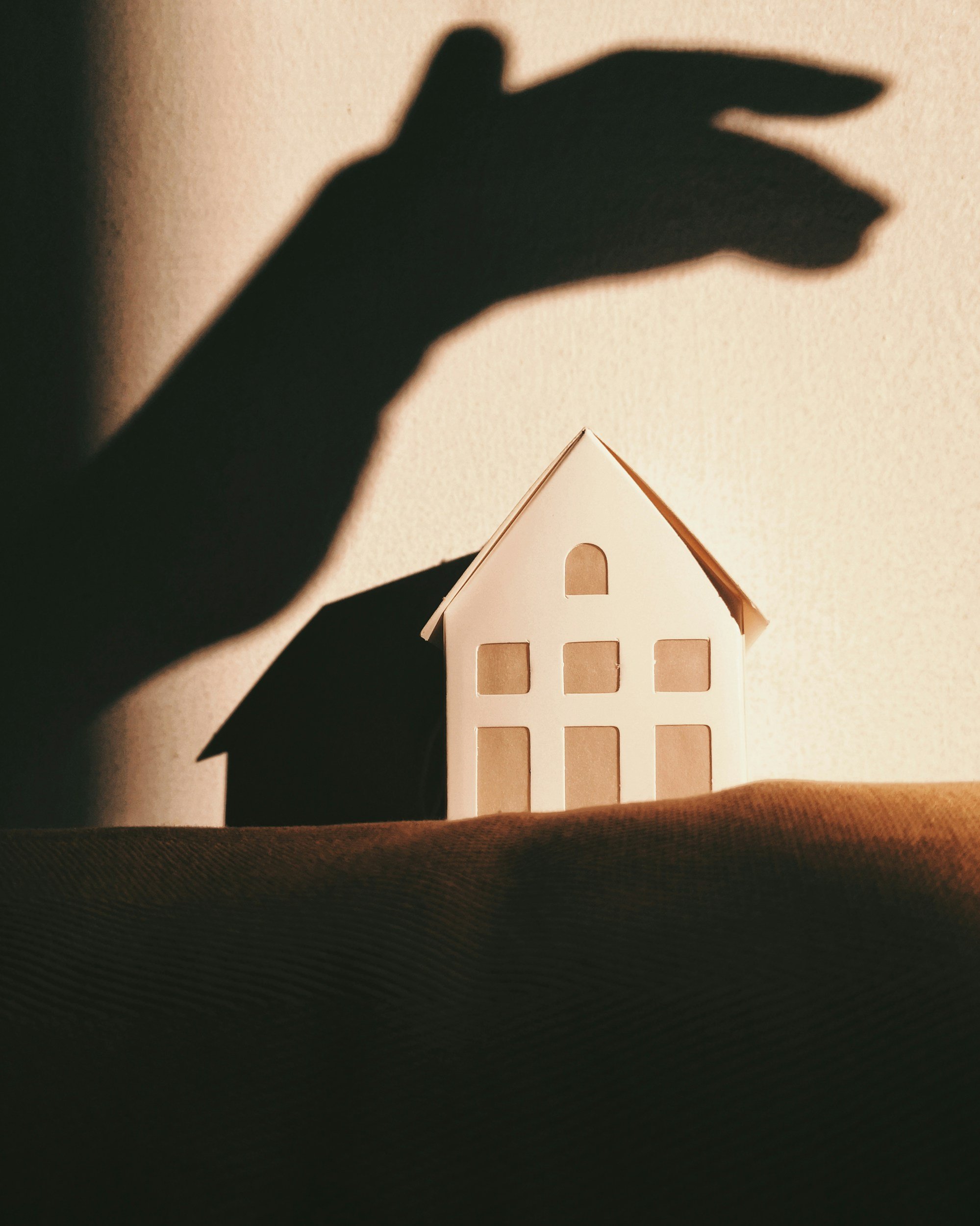 A shadow of a hand with fingers shaping a roof over a small paper house on a dark surface with a background wall.