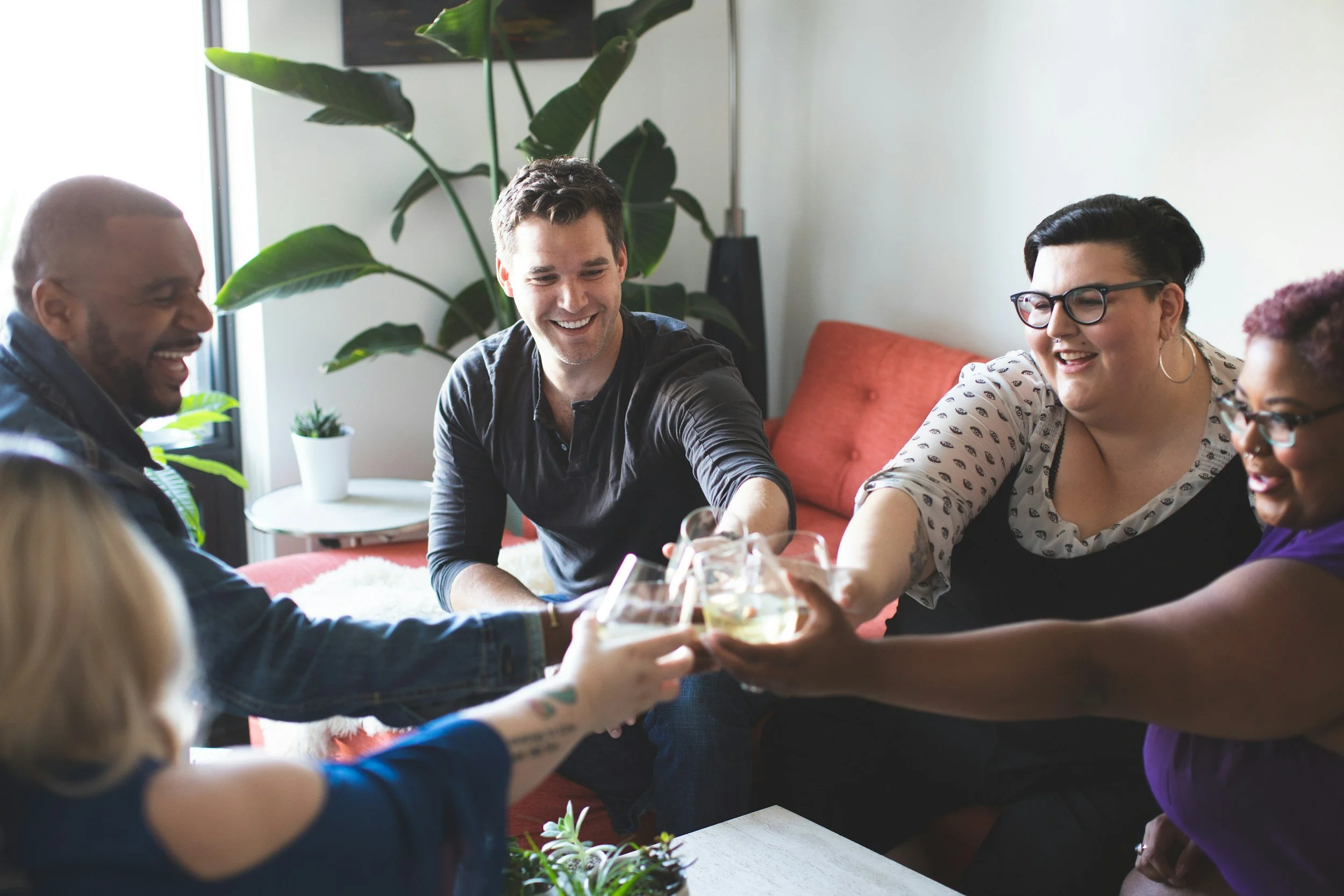 A group of diverse friends clinking glasses filled with white wine during a celebration or gathering in a cozy living room with large green plant and a red sofa.