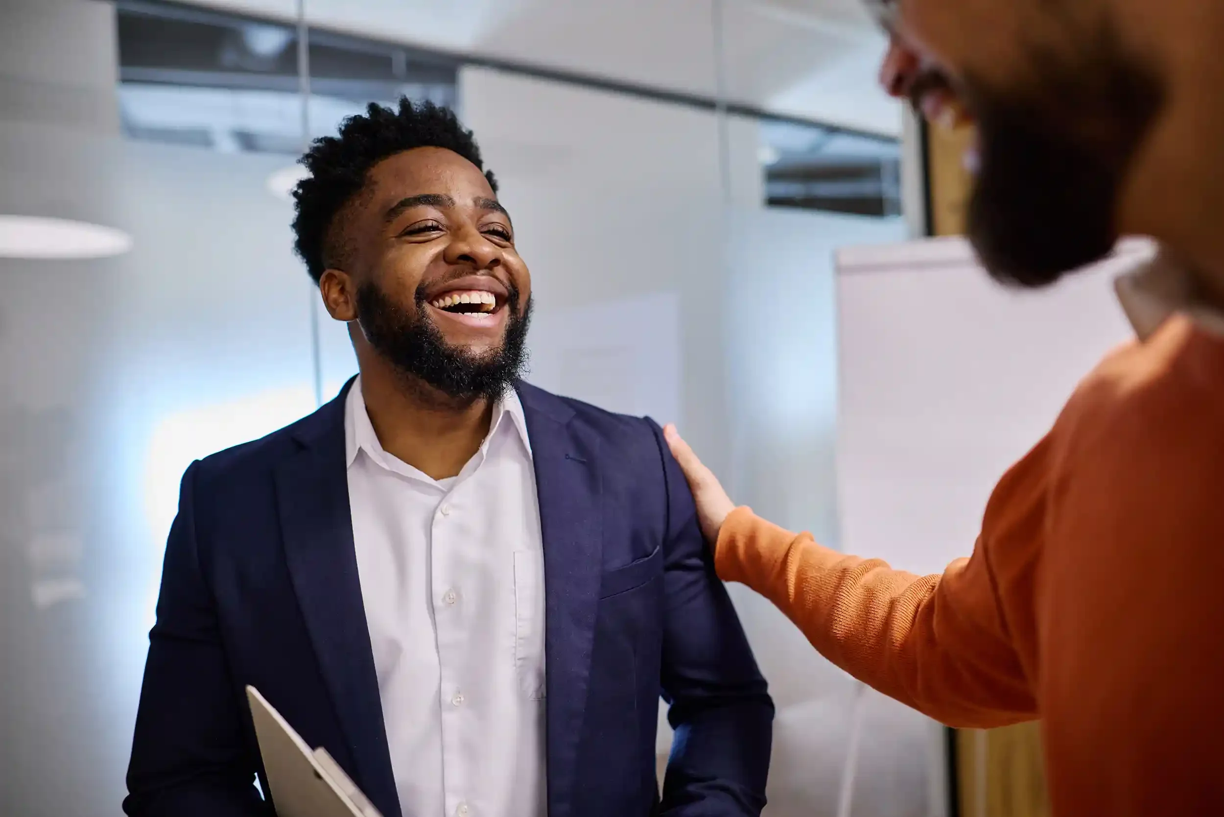 Two men smiling and greeting each other in an office setting, one with a beard wearing a navy blazer and white shirt, the other partially visible with an orange sweater