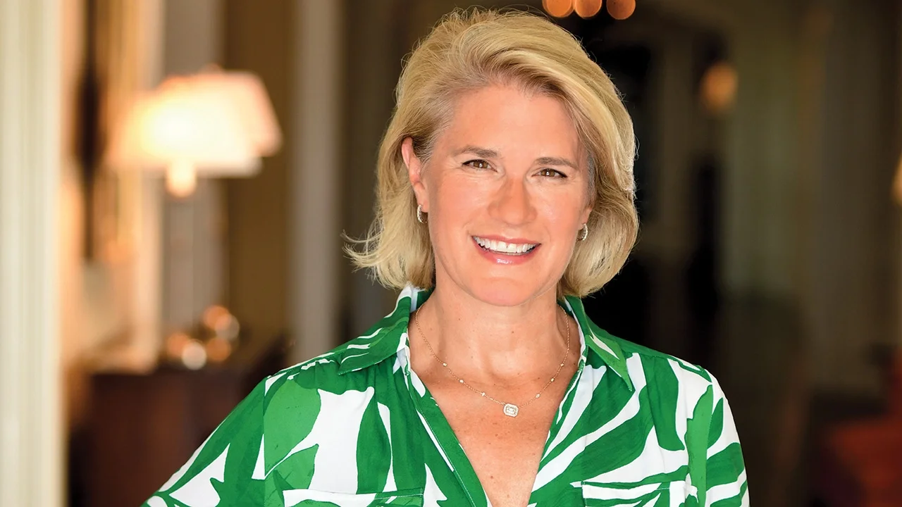 Ivy Horan, gift consultant, smiling, wearing a green-and-white patterned blouse, standing in a warmly lit home interior