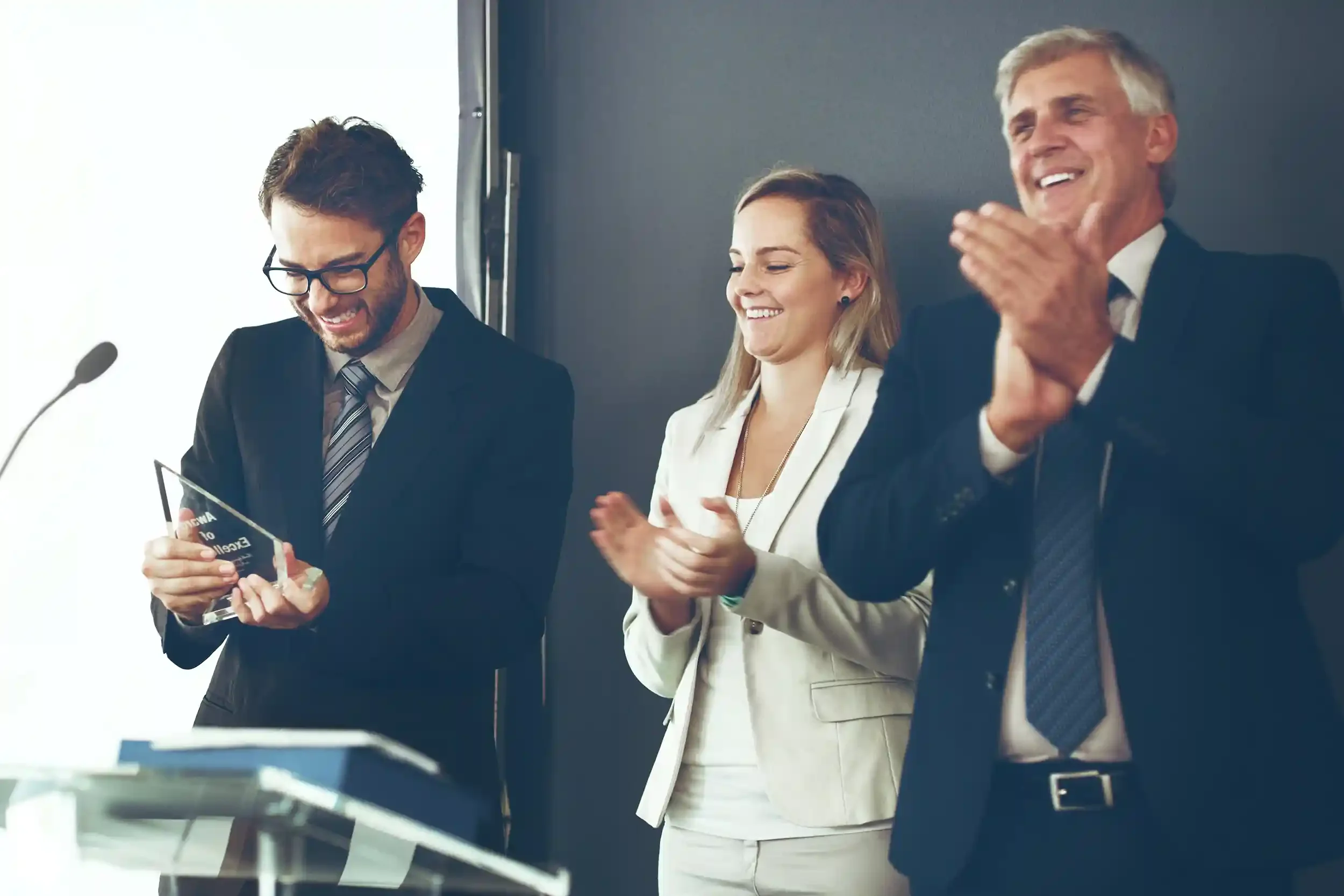 Three businesspeople in formal attire celebrating during an award ceremony or conference, with one man holding a glass crystal award and the others clapping and smiling