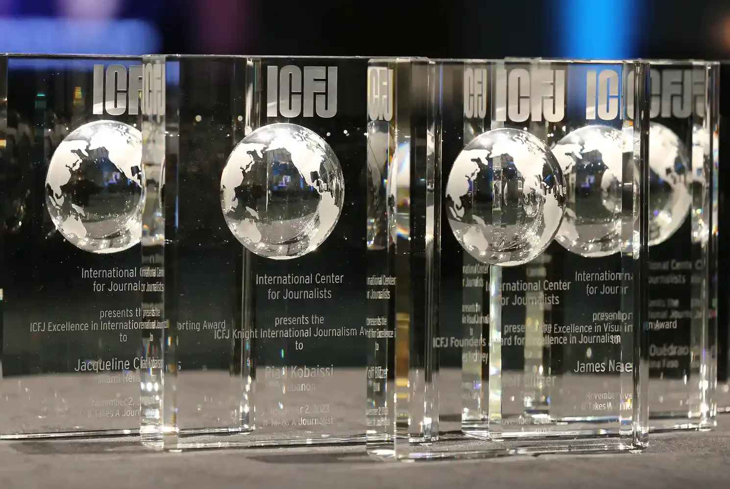 Multiple glass crystal awards with a globe engraving, inscribed with 'International Center for Journalists' and text about an excellence award in visual journalism, arranged on a surface