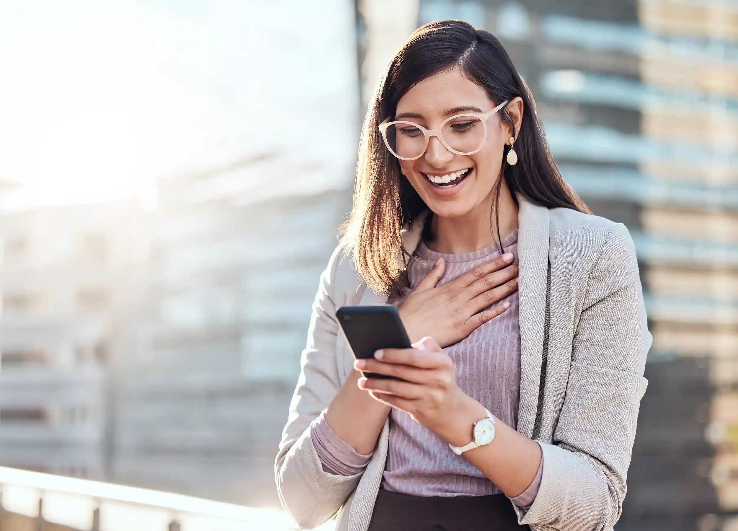 A business woman with dark brown hair and glasses, smiling while looking at her smartphone outdoors in a city environment