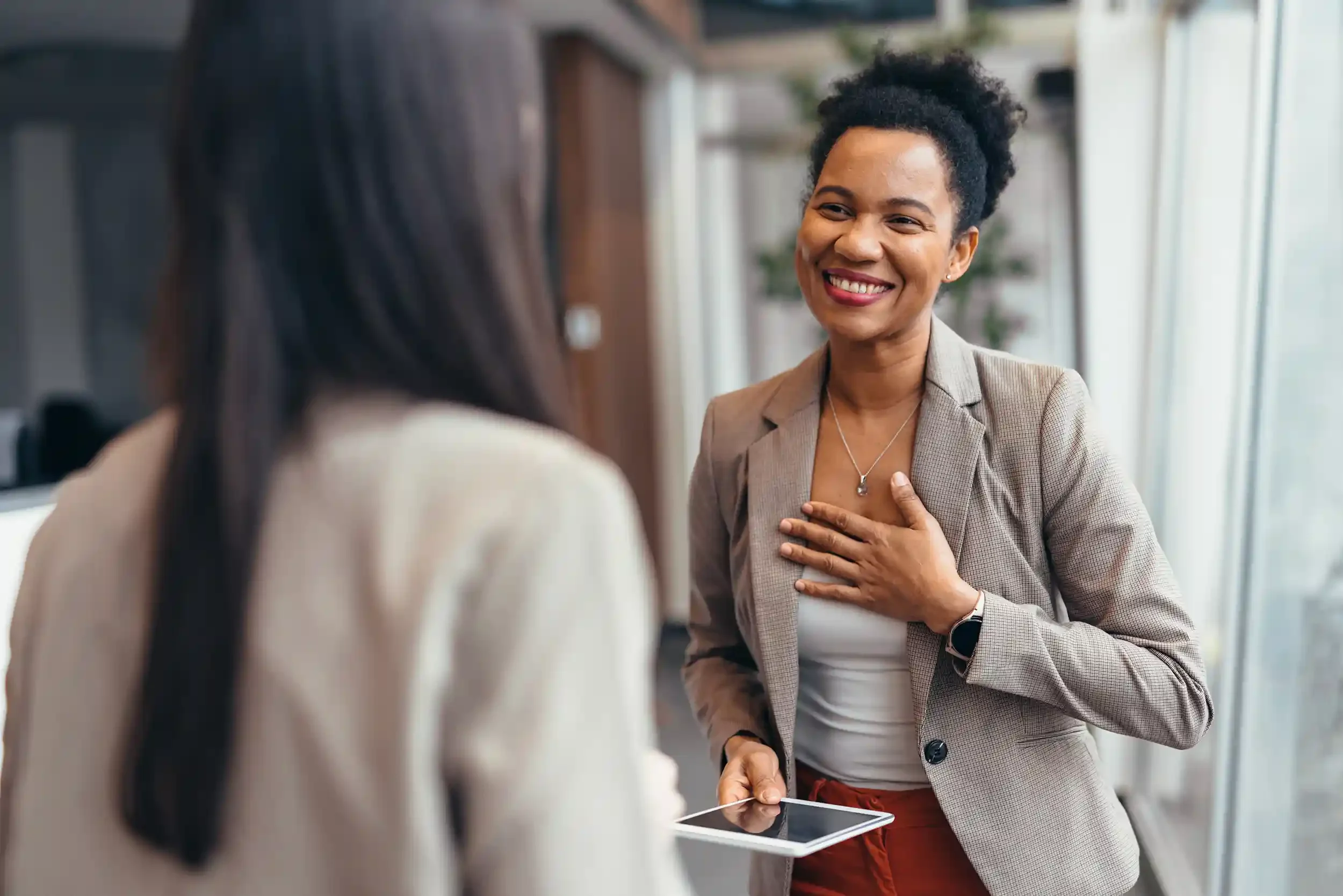 Two women having a conversation in an office setting, one woman smiling and touching her chest, holding a tablet in her other hand